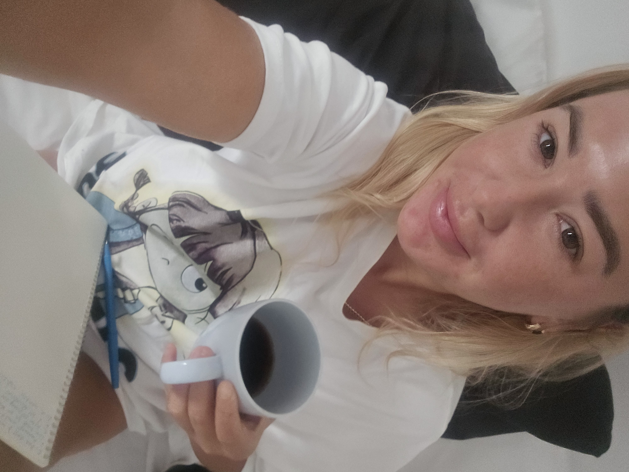 A blonde woman with fair skin and noticeable blemishes on her cheeks is shown in a close-up selfie. She is wearing a white t-shirt with a cartoon character printed on it. Her right hand holds a white mug filled with dark liquid, presumably coffee. In front of her, a white notebook and a blue pen are visible. The background appears to be a bed with white and black pillows. The woman is looking directly at the camera with a slight smile. Her hair is shoulder-length and blonde, and she is wearing a small gold hoop earring in her visible left ear. The lighting suggests an indoor setting.