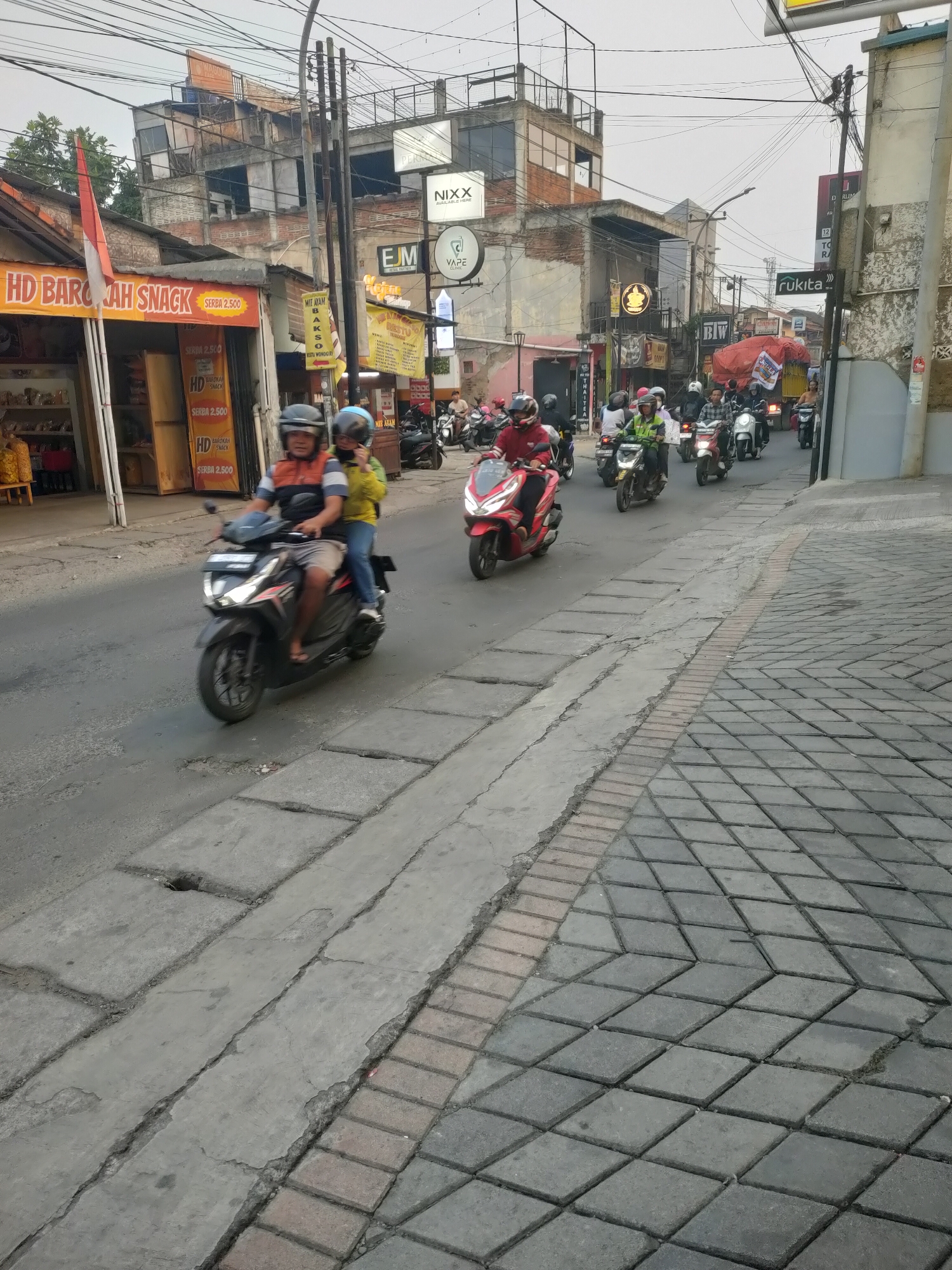 The image depicts a street scene in Cimahi, Indonesia, likely during the day. A procession of motorcyclists is traveling down a paved road, with buildings lining the left side of the street. The buildings are varied in style and condition, with some appearing older and others more modern. Signage on the buildings indicates various businesses, including "HD BAROKAH SNACK," "NIXX," "FJM," and "VAPE CLINIC." The road itself is in moderate condition, with some cracks and potholes visible. The sidewalk to the right of the road is paved with square tiles. The sky is overcast.