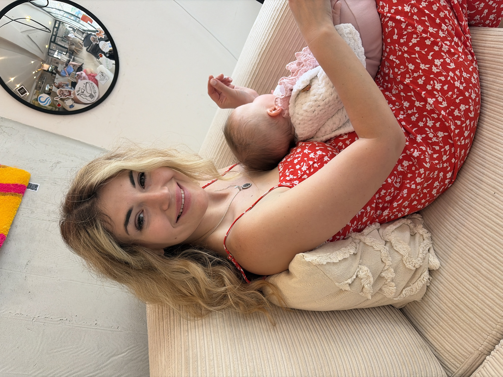 A woman is lying on a light beige corduroy sofa, cradling a baby in her arms. She is wearing a red dress with a small white floral pattern. Her hair is blonde and styled in loose waves. She is smiling at the camera. The baby is wearing a pink onesie and a white knitted sweater. A pillow is beneath her, providing additional support.
In the background, on the wall, is a round mirror with stickers affixed to its surface, reflecting the interior of a room. The mirror and rug indicate that the location is likely indoors. While I do not have enough information to pinpoint the exact location in Budva, Montenegro, the style and details suggest an interior setting, such as a home or possibly a boutique.