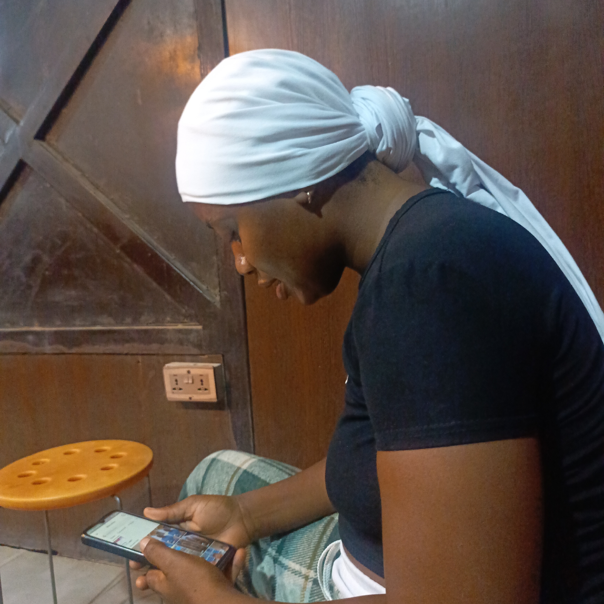 A person with dark skin wearing a black t-shirt and a white head covering is sitting, looking at a smartphone. The person is in an indoor location, next to a wooden door or wall with a light-colored electrical outlet. A small, round, orange table is nearby. The location is in the city of Unknown, Unknown.