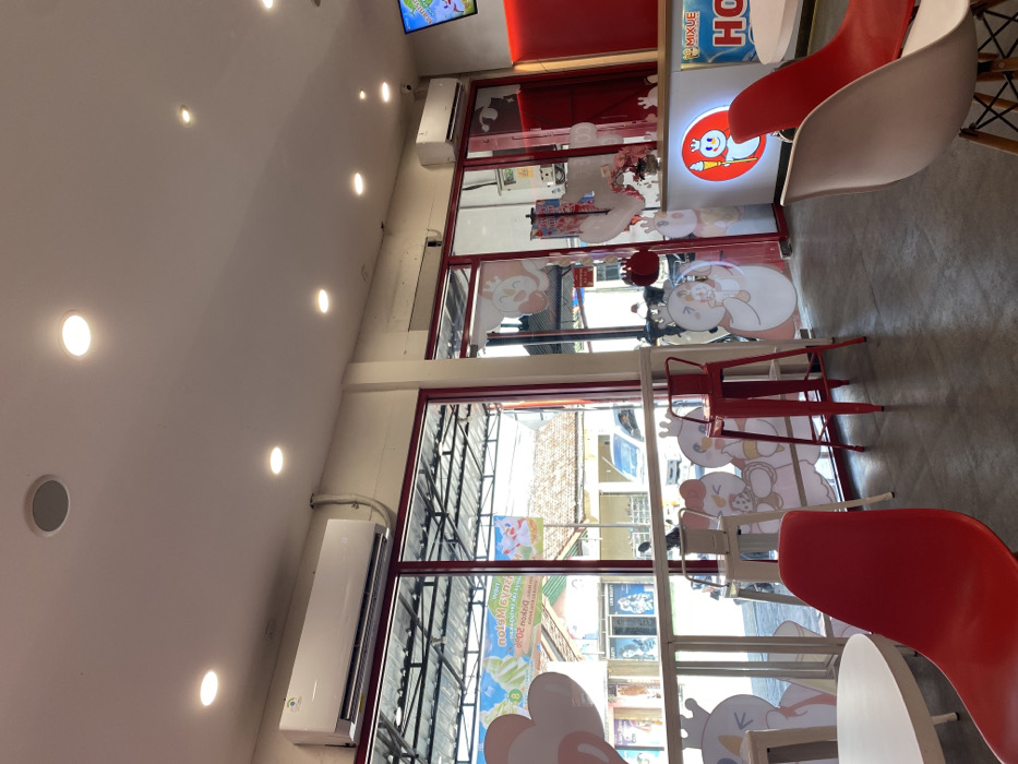 The image shows the interior of a restaurant or cafe in Kecamatan Piyungan, Indonesia (latitude: -7.837, longitude: 110.474). The cafe has a modern design with white walls, a tiled floor, and large windows that offer a view of the street outside. The ceiling has several recessed light fixtures, and a single air conditioner unit is mounted on the wall.
There are several red metal chairs and white tables placed throughout the cafe. The walls are adorned with colorful cartoon-like characters, some of which are holding ice cream cones. Behind the counter, a large sign reads "Mixue HQ," indicating the cafe's brand affiliation. The scene appears to be empty of patrons or staff, suggesting it may be closed or captured during a non-peak period.
