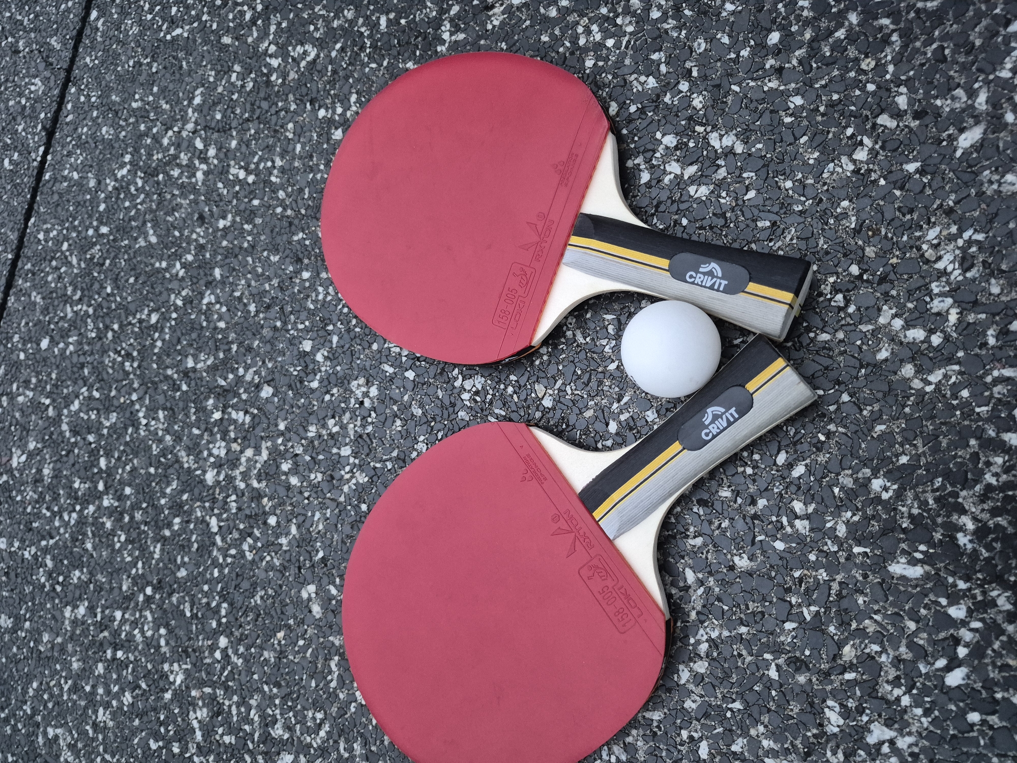 Two red-faced ping pong paddles and a white ping pong ball lie on a dark asphalt surface with white and gray flecks. The paddles are positioned with their heads facing upwards and outwards, forming a slight V-shape. The handle of the left paddle is visible on the left side of the image, and the handle of the right paddle is visible on the right. Both paddles have black and yellow accents on their handles, and one paddle has the brand name "CRIVIT" visible. The ping pong ball is located between the two paddles.