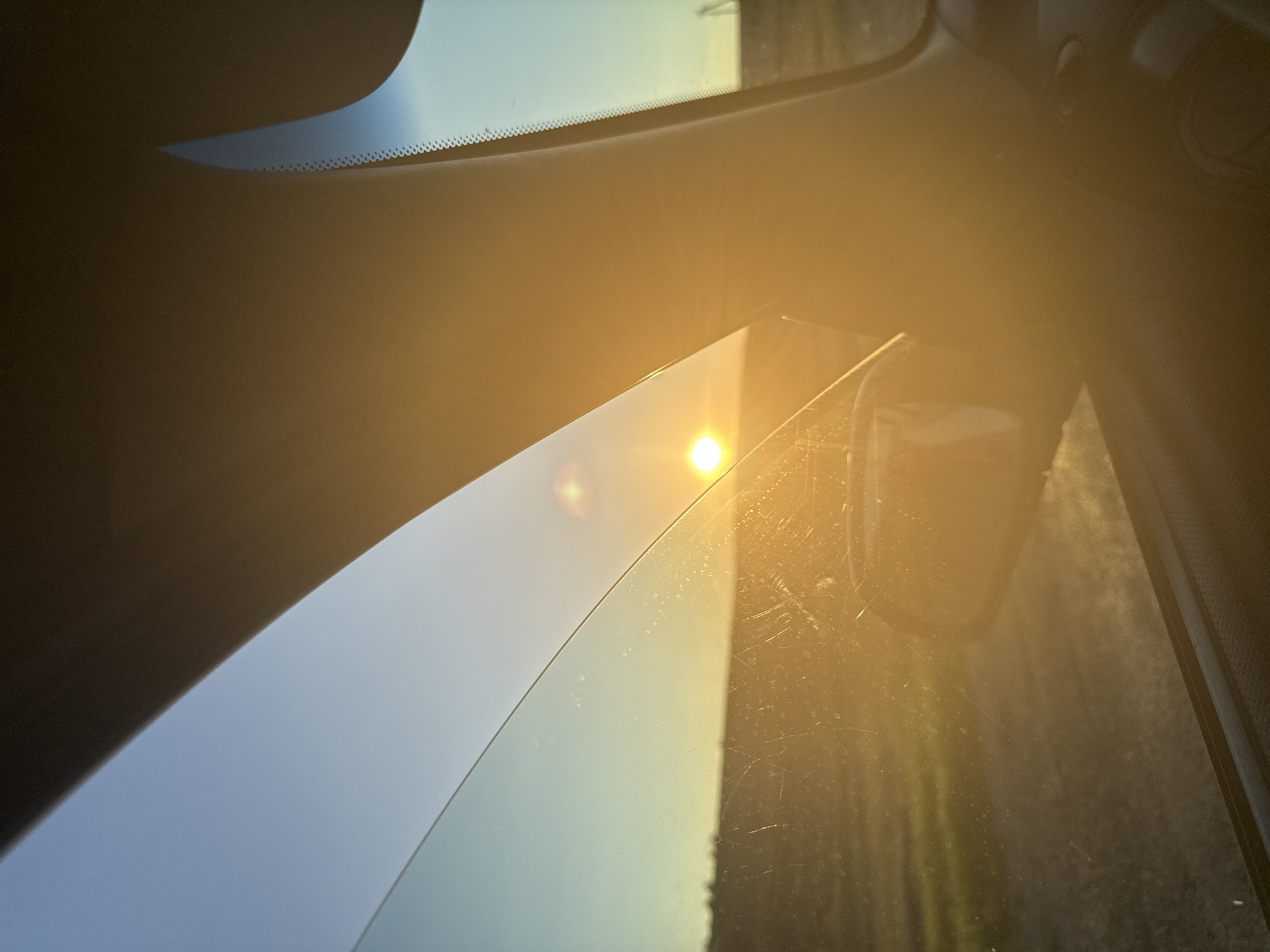 The image is a photograph taken from inside a vehicle, looking out the side window. The sun is low in the sky, casting a warm, golden glow across the scene. It appears to be either sunrise or sunset. The window glass is slightly smudged and scratched.
Through the window, a rural landscape is visible. A field stretches out, with some sparse vegetation and what looks like a dirt road or path. In the distance, a faint line of trees or structures can be discerned. The sky is a clear gradient of blue and yellow, transitioning from a pale blue at the top to a bright yellow where it meets the horizon.
The interior of the car is partially visible, including a portion of the dashboard and a side mirror. The prominent pillar of the car's interior frames the view, obscuring a portion of the landscape and sky.
The specific location in Iordanovca, Moldova, cannot be determined from the visual information presented in the image alone. The scene depicts a generic rural setting consistent with many such locations. There are no discernible landmarks or identifying features that would pinpoint the exact location within Iordanovca.