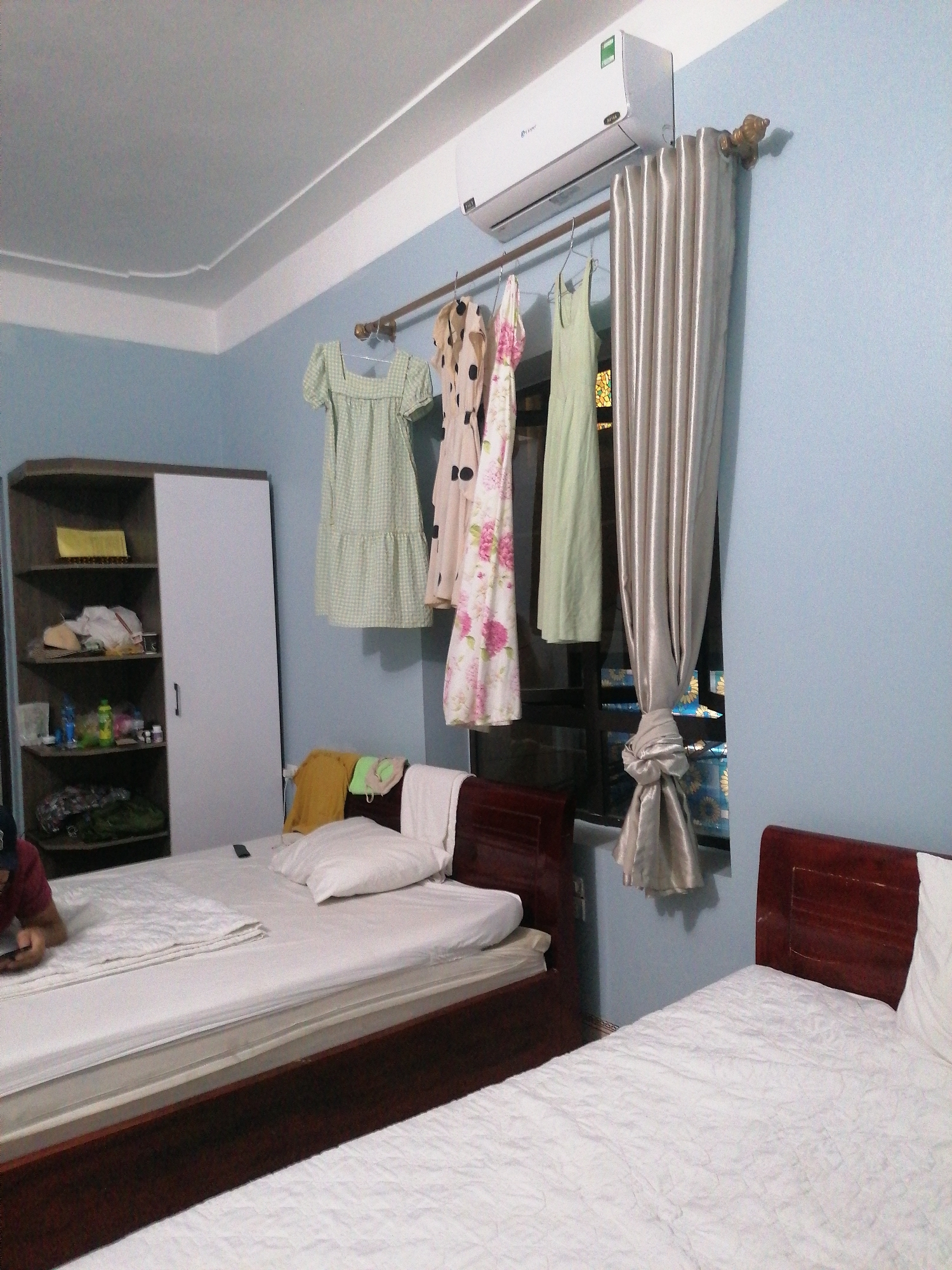 The image captures a brightly lit bedroom or guest room, likely in Bai Chay Ward, Vietnam. The walls are painted a light blue, contrasting with a white ceiling featuring decorative molding.
In the foreground, two beds with wooden headboards and white quilted bedding are visible. On the bed to the left, a person is partially visible, wearing a red shirt and dark cap, looking down at a phone or tablet. A remote control rests on the white comforter. The other bed on the right is empty except for a white pillow and some folded clothes (a yellow t-shirt, a green cloth, and a white towel) placed near its headboard.
Against the back wall, a tall, narrow cupboard with a dark wooden frame and a white sliding door stands to the left. Its open shelves are filled with various items, including folded clothes, plastic bottles, and bags. Above the window, an air conditioner is mounted, displaying the "Gree" logo, "R410A," and a green energy efficiency sticker.
A metal curtain rod extends across the window, holding several items of clothing on hangers, including a light green gingham dress, a polka-dot dress, a floral dress, and a solid light green dress. A silvery-beige curtain, tied in the middle, covers part of the window. Through the gaps in the curtain and above the clothes, decorative window elements with blue backgrounds and yellow floral patterns are visible, suggesting either a patterned window film or a view of a colorful building detail outside.
The room appears tidy, though lived-in, with items stored on shelves and clothes hanging to dry or be aired out. The lighting suggests it is daytime.