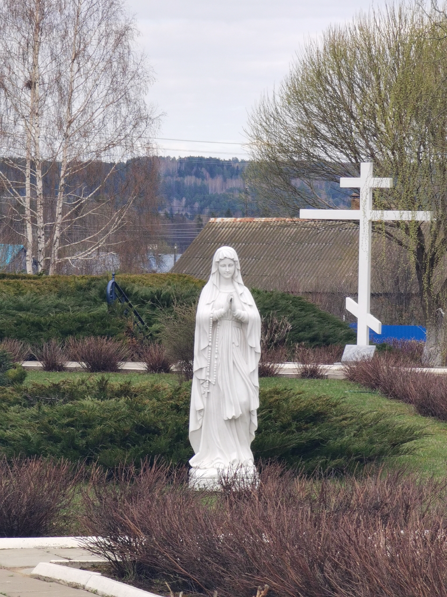 A full shot of a white statue of a woman with a rosary standing in a garden. The statue is the main focal point and is surrounded by green bushes and dry, brown shrubs. A white Orthodox cross stands behind the statue. The background includes a building with a gray roof, trees with budding leaves, and a distant forested area under an overcast sky. The scene depicts a religious monument in what appears to be a well-maintained outdoor setting. The location is Кварса, Russia.