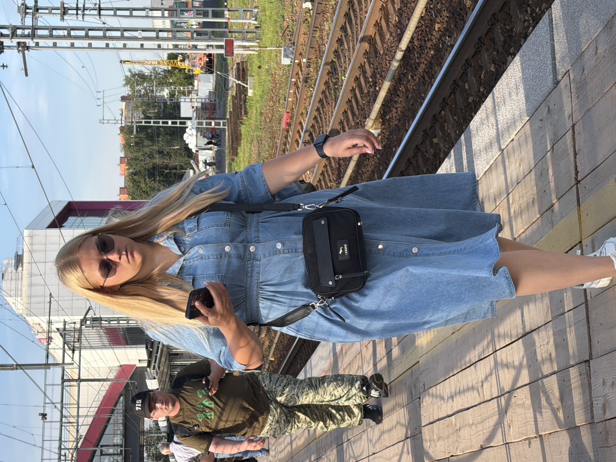A blonde woman wearing a denim dress and sunglasses walks on a wooden train platform in Shcherbinka, Russia. She holds a smartphone in her right hand and wears a black crossbody bag. To her left, a man wearing a dark green t-shirt with green graphics and camouflage pants walks in the same direction, also holding a smartphone. The train tracks are visible to the right of the platform, with overhead power lines and a distant building with a red and white design. The lighting suggests it is daytime, likely late afternoon due to the long shadows cast on the platform.