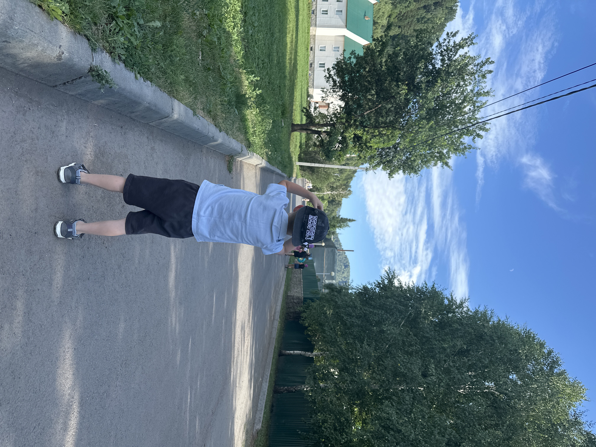 The image depicts a young boy wearing a light blue shirt and dark shorts, standing on an asphalt road and looking away from the camera. He is wearing a dark baseball cap with the word "RESPECT" printed on it. The boy is positioned in the foreground, facing down a road that curves to the right.
To the left of the road, there is a grassy area with a concrete curb and a row of trees. In the background, a white building with a green roof is visible, surrounded by trees and hills. On the right side of the road, there is a dark green metal fence, followed by more trees and a building. The sky is blue with scattered white clouds, and a crescent moon is visible in the upper left portion of the image.
The location is identified as Danilovka, Altai Krai, Russia, suggesting a rural or semi-rural setting with natural elements and some residential or communal buildings. The overall scene appears to be a sunny day.