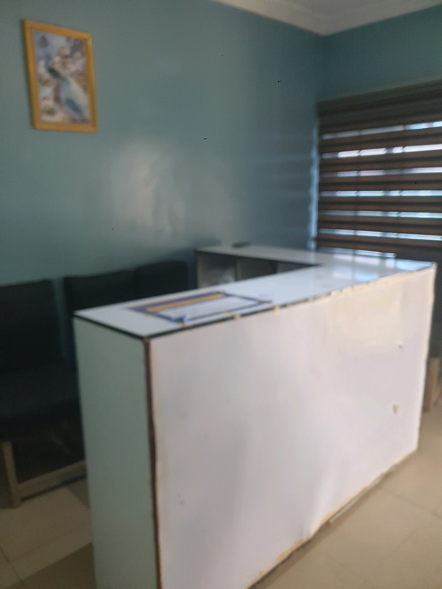 The image shows an indoor scene, likely a reception area, in Mangu Town, Nigeria. A white reception desk with a curved front is centrally positioned, its surface displaying some papers and a ruler. To the left of the desk, there are dark-colored chairs, suggesting a waiting area. The wall behind the desk is painted a light blue-green, and a framed picture, appearing to depict a bird, hangs on this wall. To the right of the desk, a window with horizontal blinds is visible. The overall environment appears to be clean and organized, with neutral flooring.