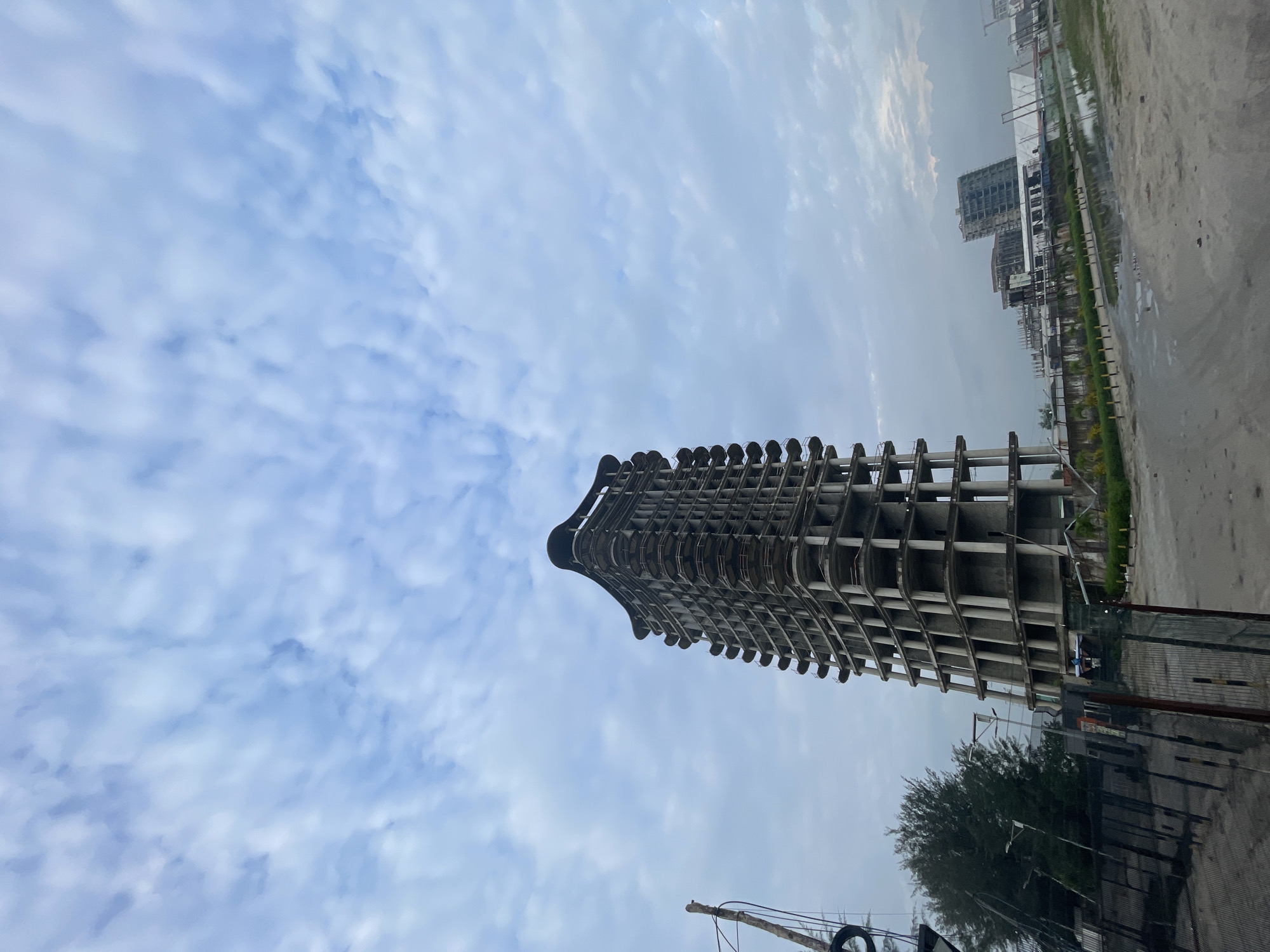 An unfinished, tall, concrete building stands prominently in the foreground of the image. The building has a unique, curved design with multiple balconies or floor extrusions. It appears to be in a state of construction or abandonment, with exposed concrete and a lack of finishing.
To the left of the main building, a bare, weathered wooden pole is visible, possibly a utility pole or part of a structure. A dark fence runs across the bottom left of the frame, with some vegetation behind it.
In the middle ground and background, other buildings are visible, also appearing to be under construction or in a similar unfinished state. These include several multi-story structures and what looks like a long, low-lying building with scaffolding. The environment around the buildings is a mix of sandy ground, some puddles of water, and patchy greenery. The sky above is filled with a pattern of light, wispy clouds against a pale blue background.
The scene is located in the city of Ilado, Nigeria, and captures a developing or stalled construction area within the urban landscape. No people are visible in the image.