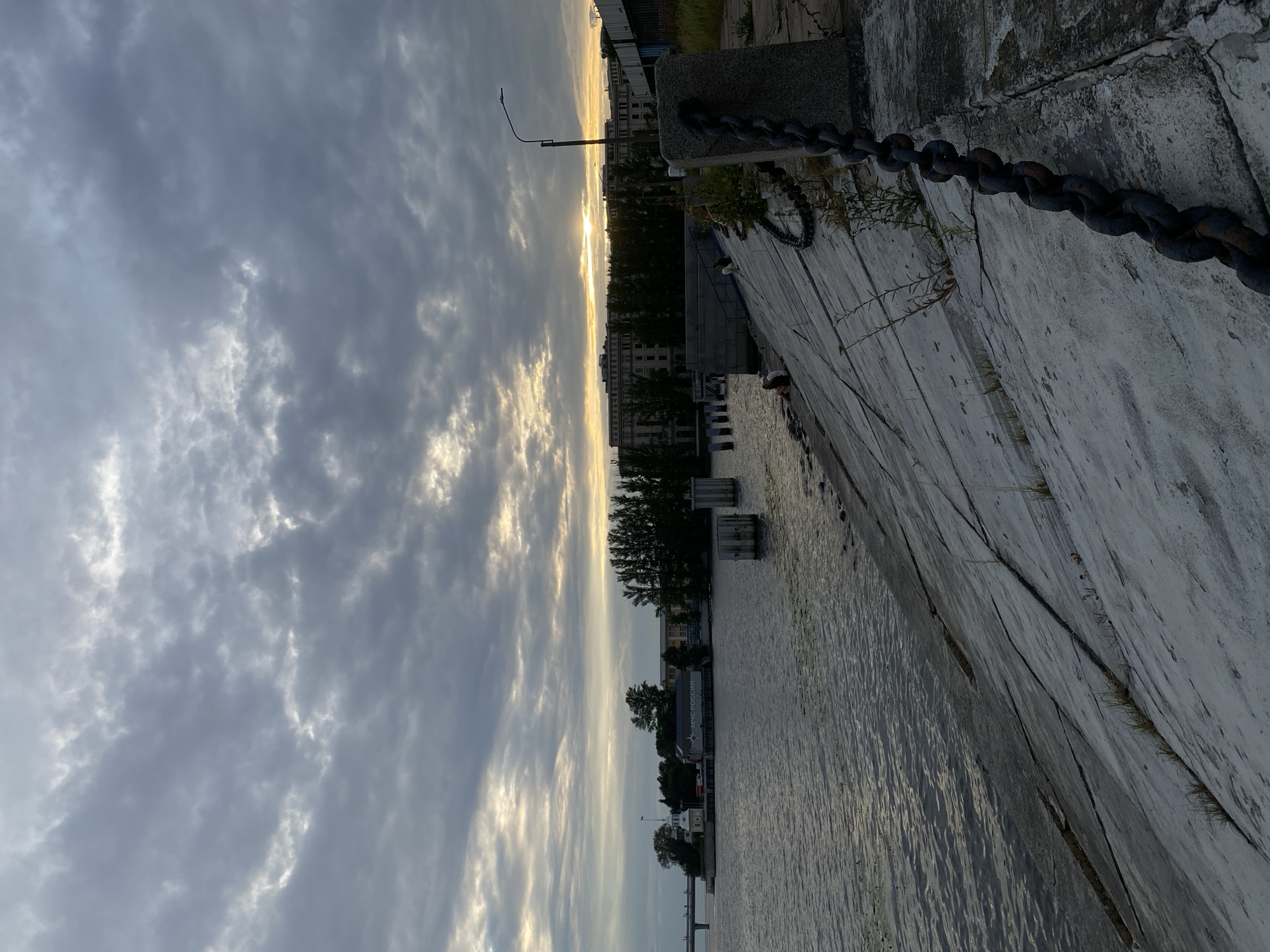 The image depicts a waterfront scene at dusk, likely in Saint Petersburg, Russia. A broad, calm body of water occupies the left side of the frame. The right side features a steeply sloping concrete embankment, reinforced with a thick, dark chain secured to a stone post and extending towards the bottom right corner. Scattered vegetation grows from cracks in the embankment. In the background, across the water, a long, multi-story building complex with a dark facade is visible, partially obscured by a line of tall trees. Further to the left, a bridge spans the water, and a smaller building with a distinctive white tower and the inscription "МЧС РОССИИ" (Ministry of Emergency Situations of Russia) stands on the shore. The sky is overcast with dramatic clouds, but a band of golden light from the setting sun breaks through near the horizon, illuminating the upper portion of the background buildings and trees. A few individuals are seated on the embankment, facing the water, with one person appearing to be in a crouching position. The overall atmosphere is tranquil, with a sense of early evening stillness.