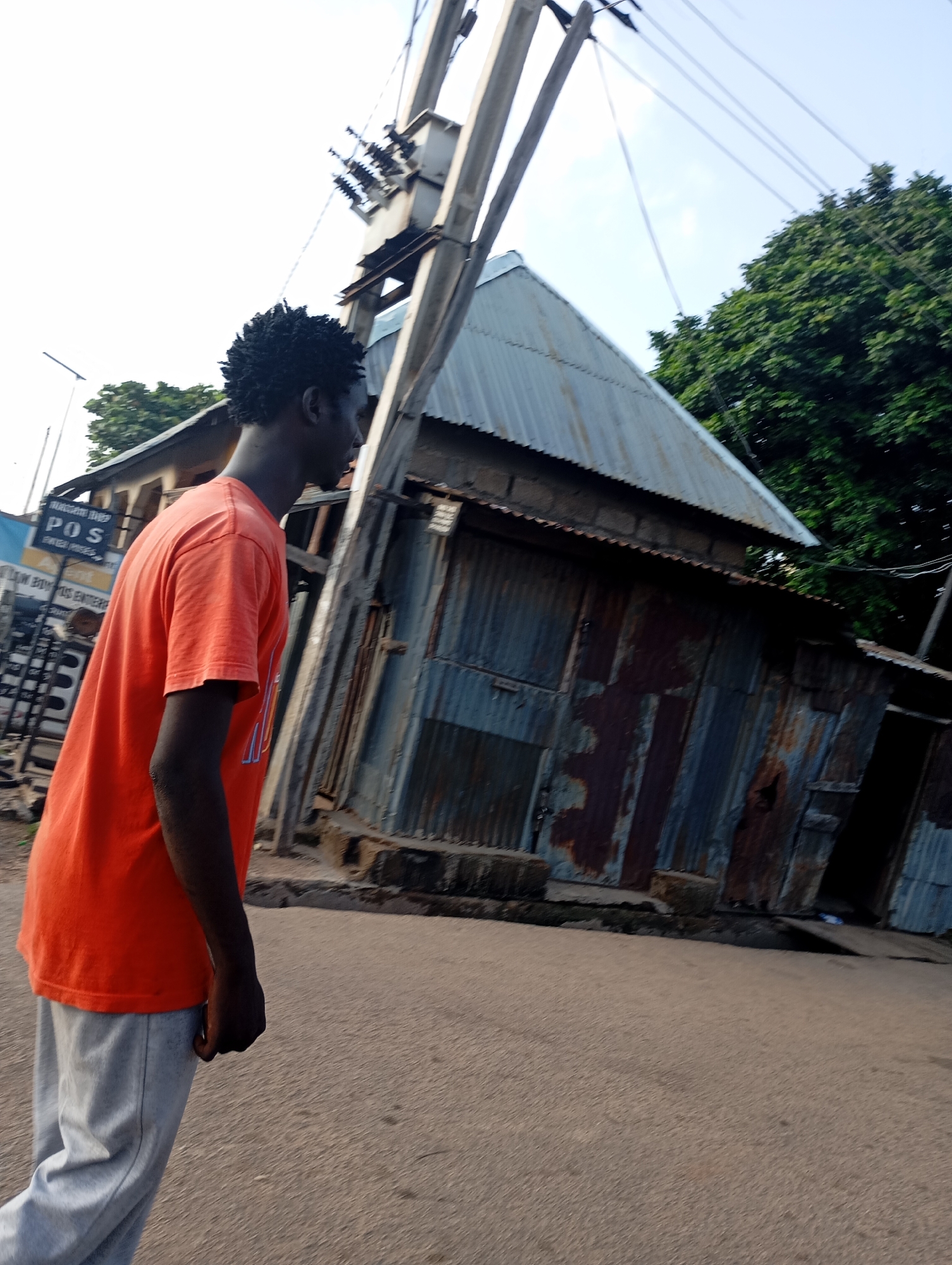 A young dark-skinned male is captured from the chest up, side profile facing right, wearing an orange t-shirt and light gray pants. He stands on an asphalt road in front of a cluster of corrugated metal buildings that appear weathered and rusted. To the left, a sign reads "POS" indicating a Point of Sale service. Behind him, a tall utility pole with electrical wires and transformers looms overhead against a bright sky. Green foliage is visible in the background. The overall scene suggests an outdoor environment in a potentially developing urban area.