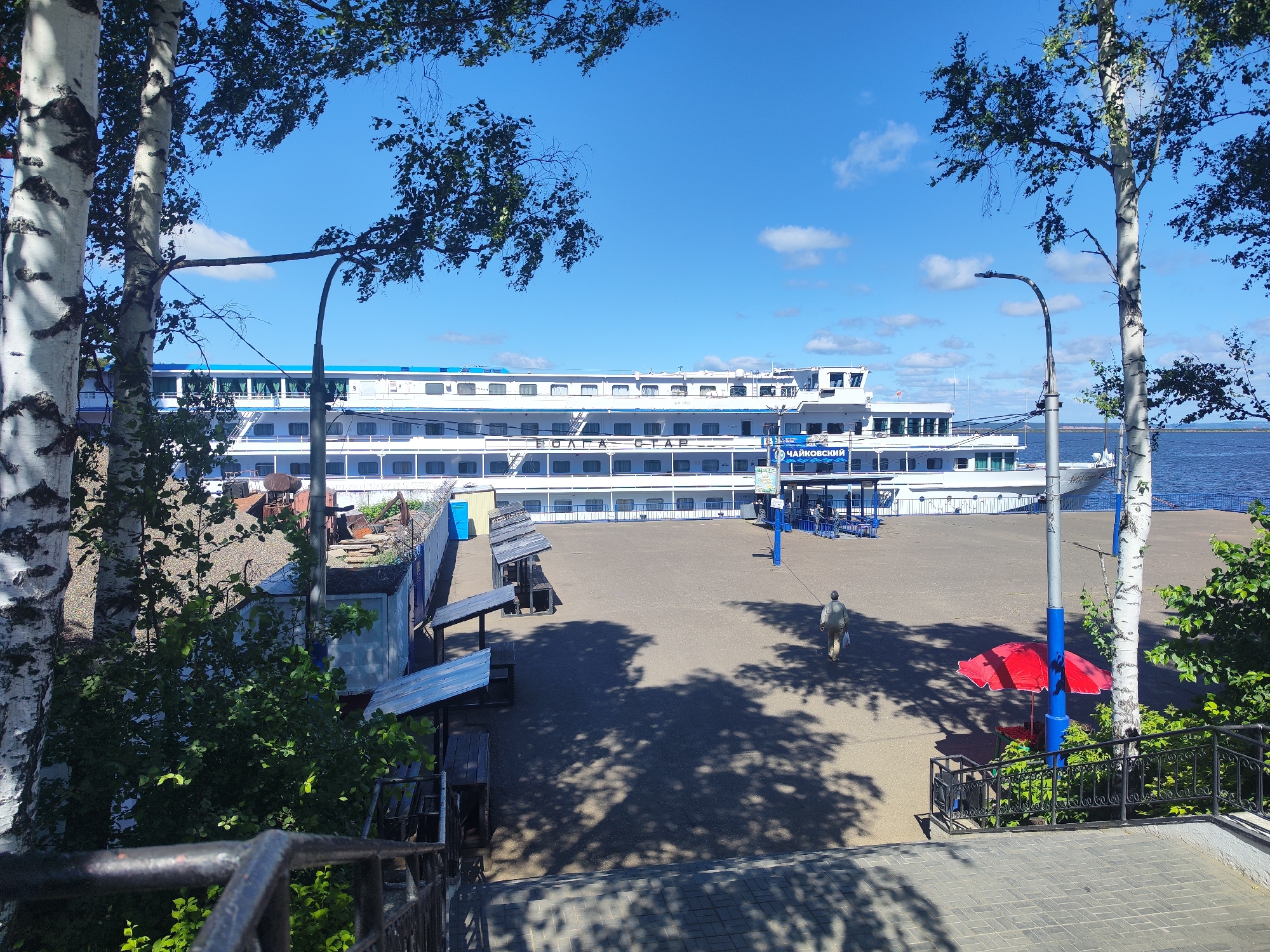 In Chaikovsky, Russia, a large white passenger ship, identified by the Cyrillic text "ВОЛГА СТАР" and "ЧАЙКОВСКИЙ" on its side, is docked at a waterfront. The ship has multiple decks with rows of windows. In front of the ship, a paved area serves as a promenade or waiting area, featuring several covered wooden benches and tables. A single person is walking away from the ship towards the right side of the frame, carrying a white bag. A red umbrella is set up near a blue pole, providing shade over a small table and chairs. The scene is framed by trees with white bark and green foliage in the foreground and middle ground, with their shadows cast on the ground. In the background, a body of water, likely a river or reservoir, stretches to the horizon under a bright blue sky with scattered white clouds. The overall impression is a sunny day at a riverside port.