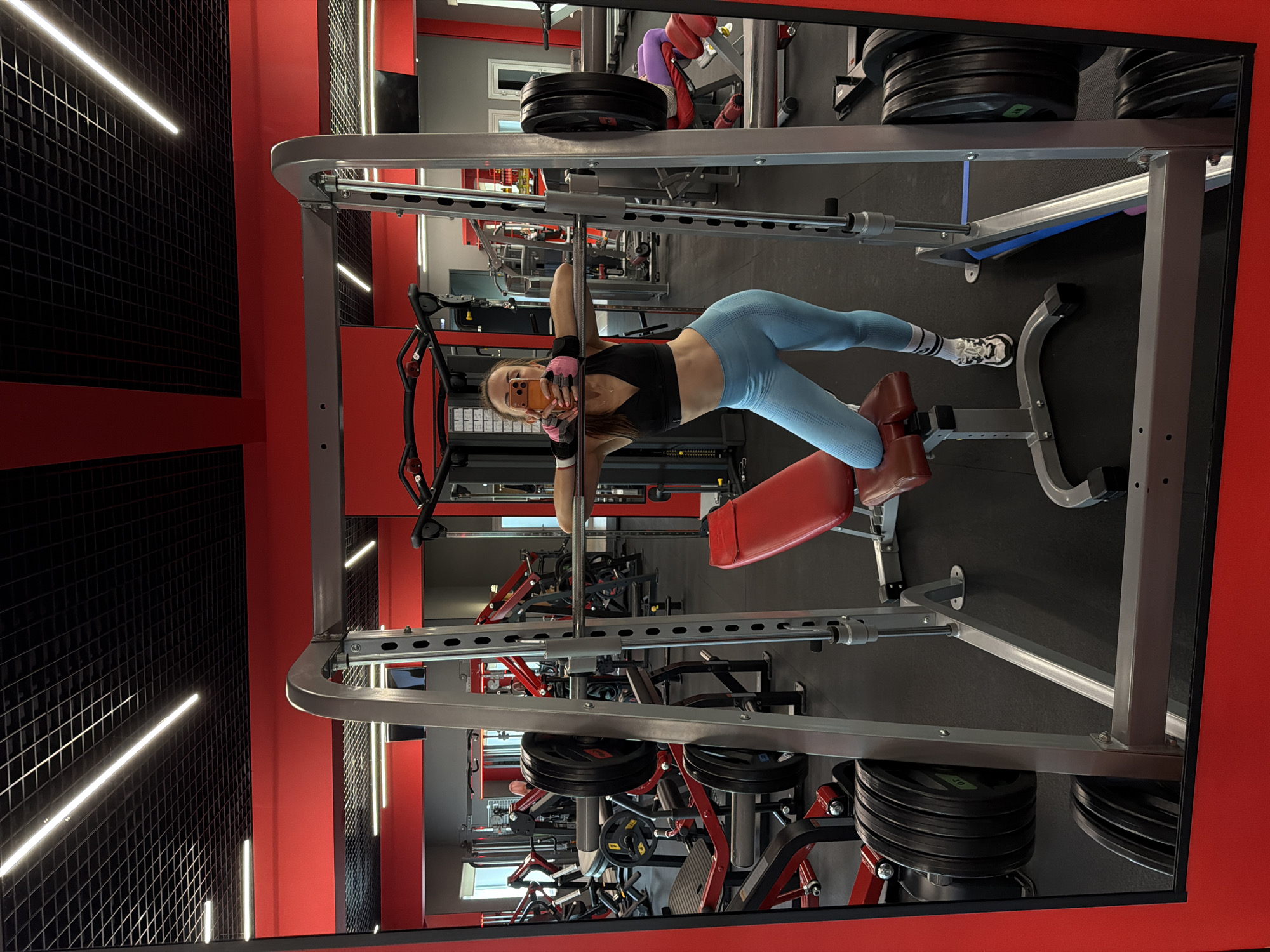 In Birobidzhan, Russia, a young woman with light brown hair, wearing a black sports bra, light blue leggings, pink gloves, and white athletic shoes with black accents, poses for a selfie in a gym. She is leaning on a barbell in a Smith machine, with her left knee bent and resting on the attached bench. The gym features red accents on its equipment and walls, with black tiled ceilings and fluorescent lighting. Other weightlifting machines and equipment are visible in the background, reflected in mirrors.