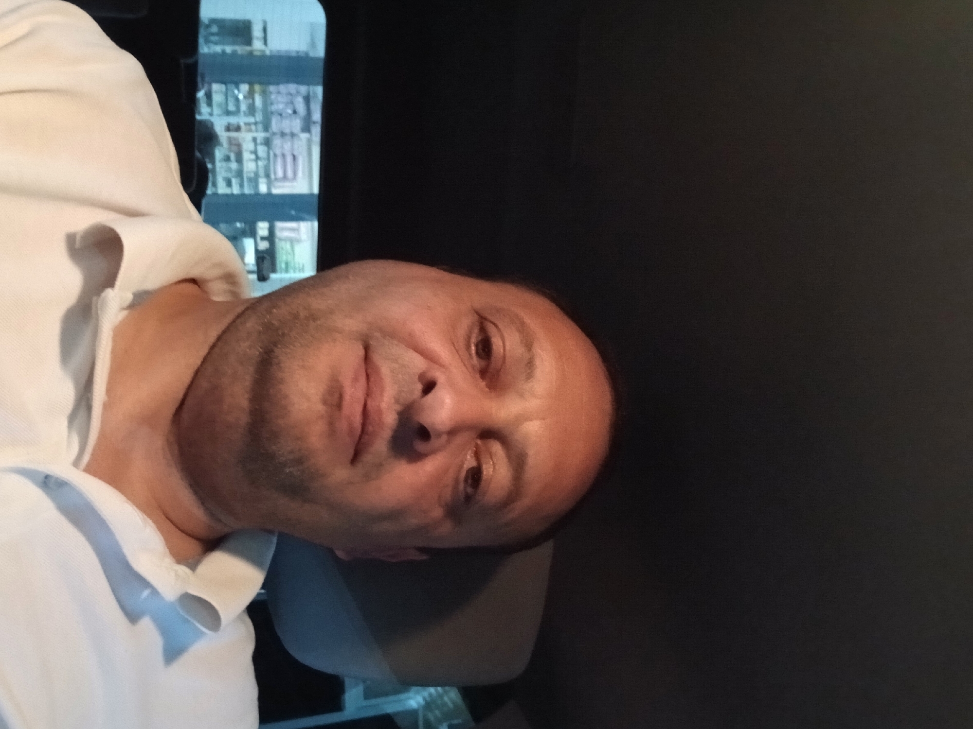 A close-up, indoor selfie shows a man in a white polo shirt. The man's head is centered in the frame, with the top of his head out of view. His face is turned slightly to the left, and he is looking directly at the camera. The background is dark, with hints of shelves filled with products visible through a window behind him. The overall lighting is dim, with some light reflecting on the man's forehead and cheek. The man appears to be in a car, given the headrest visible on the right side of the image and the dark interior of what appears to be a vehicle. The scene is a personal snapshot, possibly taken while traveling or waiting. There is no indication of the specific location in Telavi, Georgia, beyond the possibility that the man is a resident or visitor to the city.