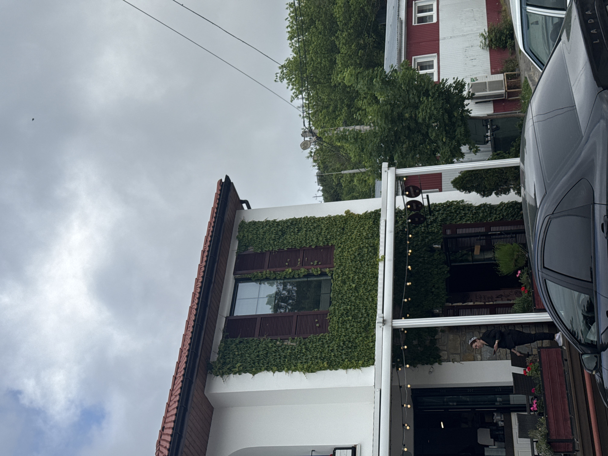 The image depicts a building in what appears to be a commercial area, likely in Abrau-Dyurso, Russia. The building is partially covered in greenery, particularly ivy, around a window with brown shutters. It has a red-tiled roof. To the right, there is an outdoor seating area with a white awning strung with lights, partially obscured by a dark-colored car. A person is visible in the seating area, dressed in dark clothing, standing near tables and chairs. A building painted in red and white and also a lot of green plants and trees can be seen in the background. The sky is cloudy.