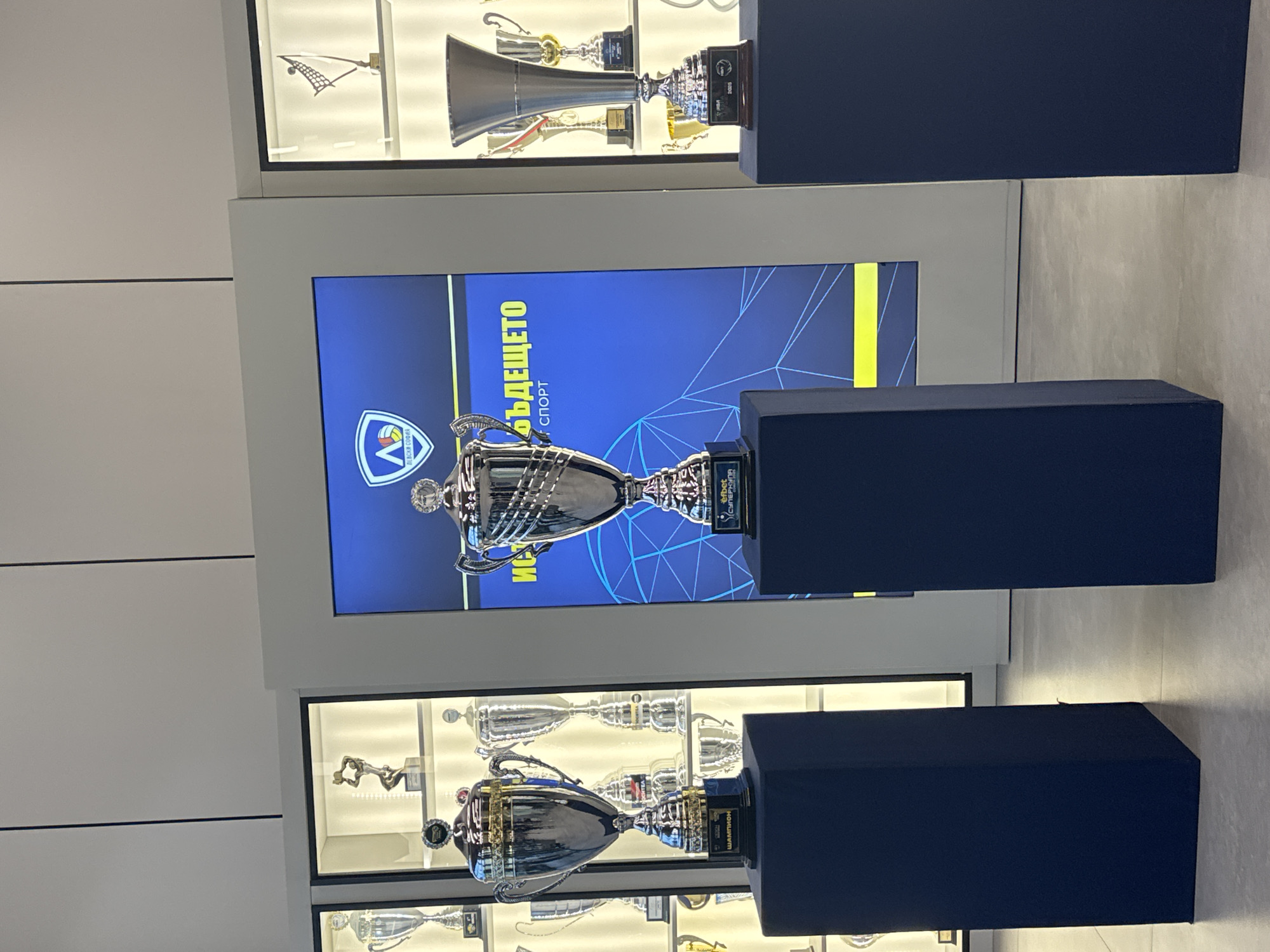 This image captures an indoor display, likely within a sports museum or club headquarters in Sofia, Bulgaria, showcasing a collection of prestigious trophies. The scene is dominated by several large, gleaming silver trophies, with two particularly prominent ones positioned on dark blue, fabric-covered pedestals in the foreground.
Between these two pedestals, a large vertical screen displays a vibrant blue graphic with yellow accents. On this screen, a trophy similar to the one on the middle pedestal is depicted, alongside a stylized logo resembling a 'V' with a volleyball inside, and the Cyrillic text "БЪДЕЩЕТО" (The Future) and "СПОРТ" (Sport). Below the logo, smaller text likely reads "УМНИТЕ ВЪЛЦИ" (The Smart Wolves), referring to a sports club or academy. Visible text on the base of the trophy displayed on the screen includes "efbet Суперлига" (efbet Superliga), suggesting a championship title.
Above and below the central screen, recessed glass display cases are filled with numerous other trophies of varying designs and metallic finishes, some silver, some golden. One trophy in the upper case resembles a large bugle or trumpet, while another in a lower case depicts a reclining figure. On the base of the lower trophy on a pedestal, the Cyrillic word "ШАМПИОН" (Champion) is visible.
The setting features light-colored walls and appears brightly illuminated by artificial lighting, creating a clean and modern aesthetic suitable for presenting significant achievements. No people are present in the image, focusing solely on the celebratory display of athletic success.