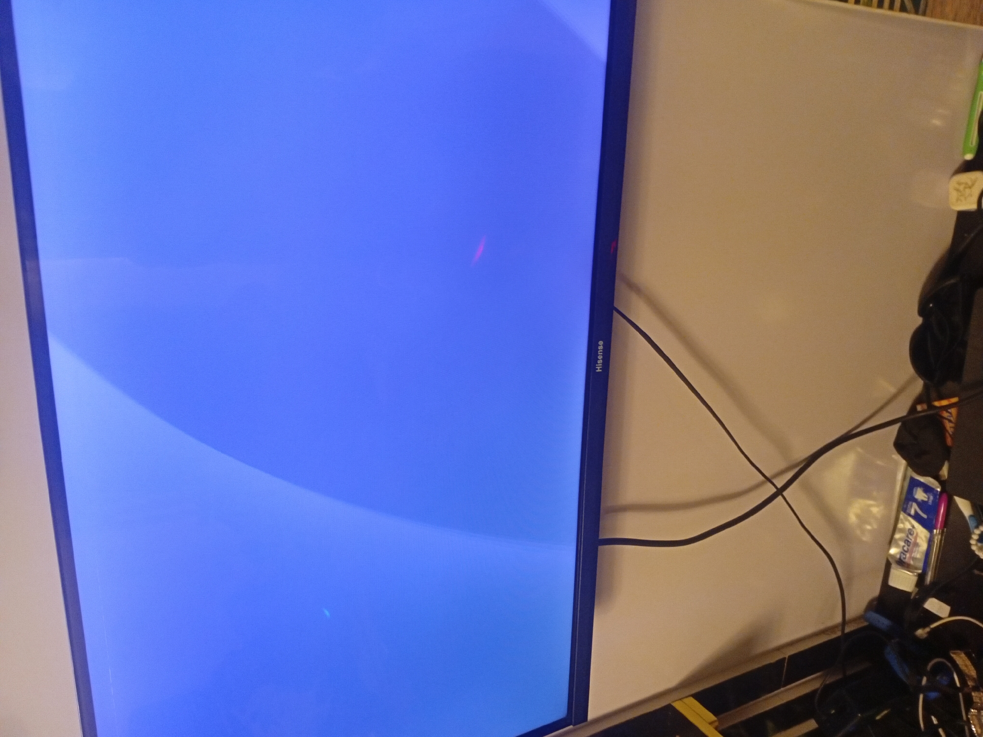 The image shows a close-up, angled view of a Hisense television screen displaying a blue image, positioned in an indoor setting. The screen is predominantly a bright blue, featuring a subtle, lighter blue or white curved pattern in the upper left and lower left sections. A distinct, glowing red streak, approximately 3-4 inches long, is visible horizontally across the screen, slightly to the right of the center.
To the right of the television, a light-colored wall, possibly beige or off-white, is visible. Several black cables are draped across this wall and the adjacent surface. On this surface, beneath the cables and to the right of the television, there is a collection of miscellaneous items. These include what appears to be a green pen or marker, a small blue and silver packet with the text "acare" and the number "7" visible, and other indistinct dark objects, possibly electronic accessories or bags. The TV itself has "Hisense" printed vertically on its right-side bezel. No human subjects or discernible activities are present. The lighting suggests an indoor environment.