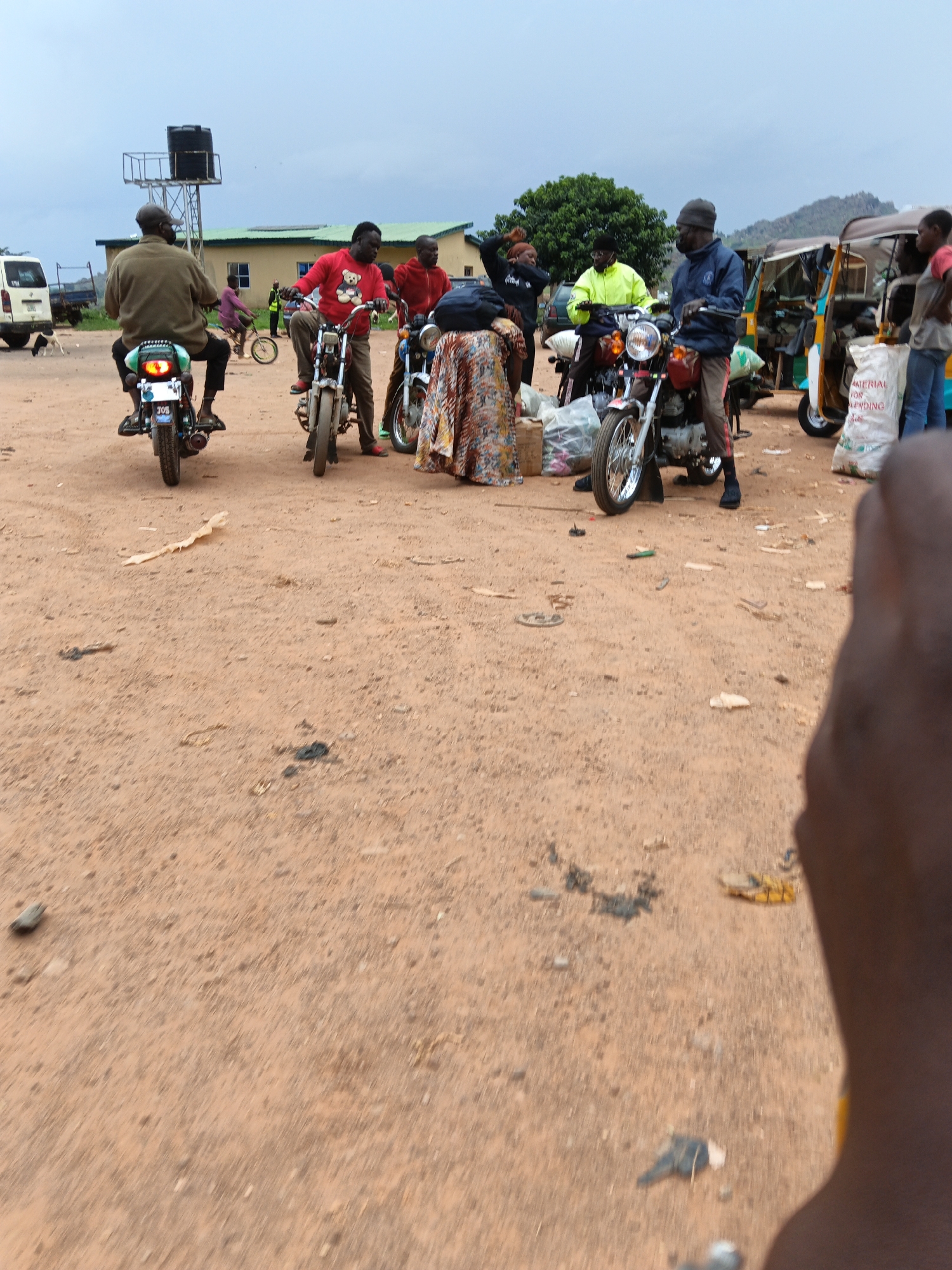 An overhead drone shot shows a dusty, uneven outdoor market scene in an indeterminate urban location. In the foreground, a hand, partially visible, looms over the scene. A man on a motorbike, seen from behind, rides away from the camera, his green jacket and cap clearly visible. He is accompanied by a white dog. Further in the background, a group of people gathers around motorbikes and a three-wheeled vehicle. One man, wearing a red sweater with a teddy bear graphic, stands next to a motorbike. Another man, in a blue jacket and beanie, sits on a motorbike. Several bags, some white and labeled, are scattered around the market. The overall setting is characterized by dirt ground, a few buildings in the distance, and a cloudy sky.