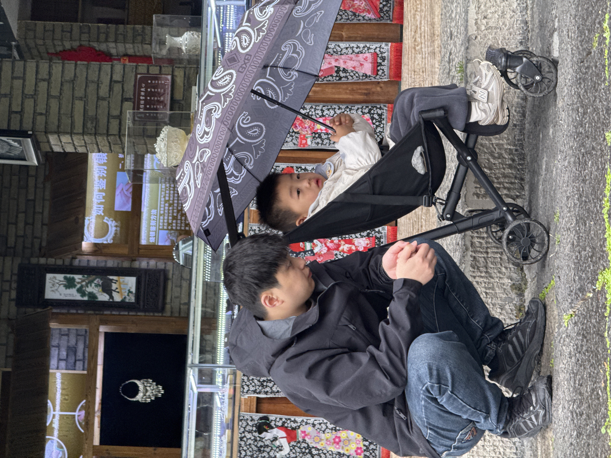 The image captures a man sitting beside a child in a stroller beneath an umbrella. The man is wearing a dark jacket over a gray hooded sweatshirt and blue jeans, with black sneakers on his feet. The child is in the stroller, covered by a gray umbrella patterned with large paisley shapes. The background includes a brick wall and glass display cases with various items. The image was taken in the city of Zhuoshui, China.