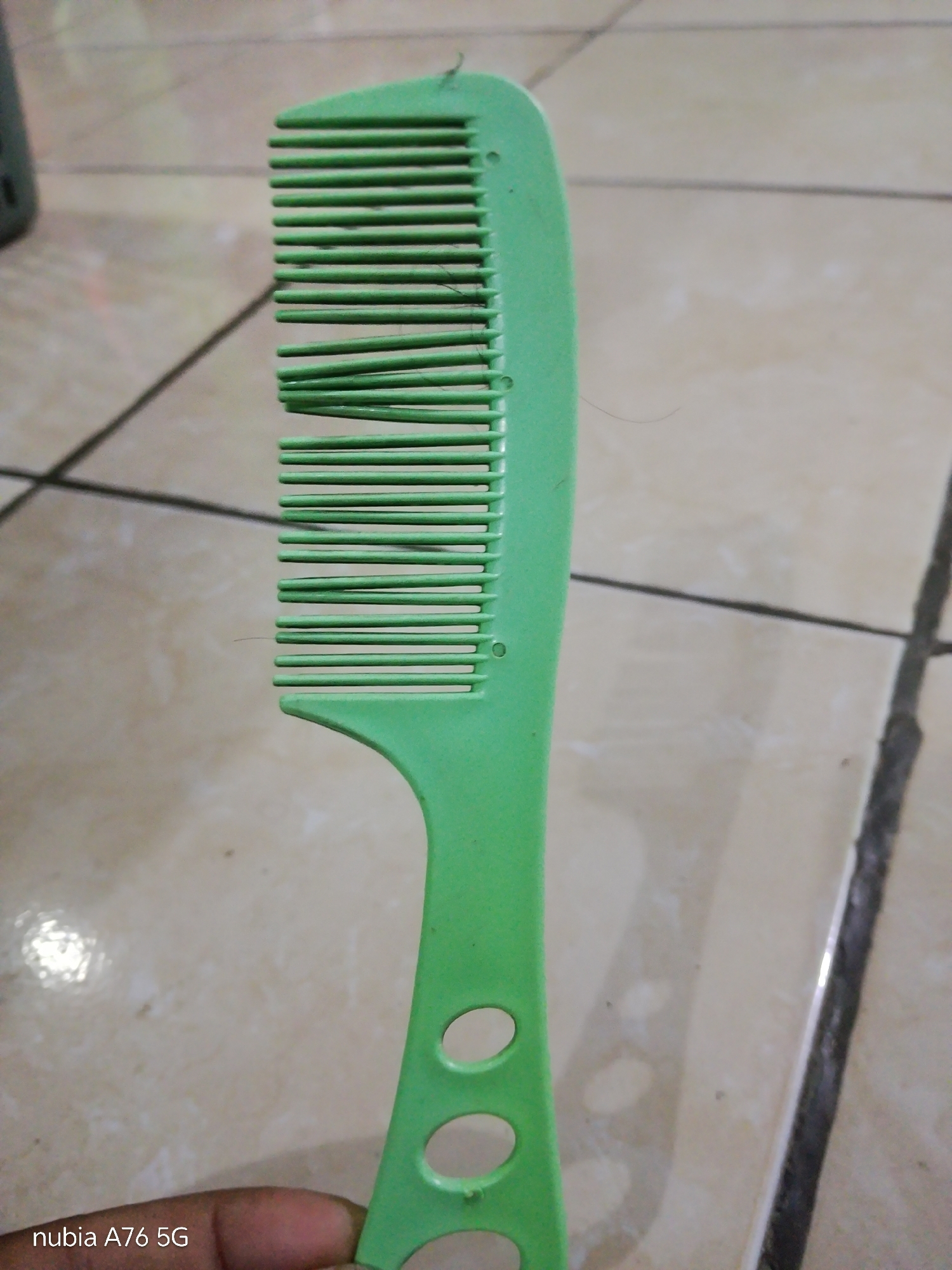 A bright green plastic comb rests on a tiled floor. The comb is held by a hand whose skin tone is dark. The tiles are a light tan color with dark grout lines. The comb has a wide handle with three circular cutouts and fine teeth. A few strands of dark hair are caught in the teeth of the comb. The setting appears to be indoors. The lower left corner of the image displays the text "nubia A76 5G", indicating the device used to capture the photograph, not a location in Binuang, Indonesia.
