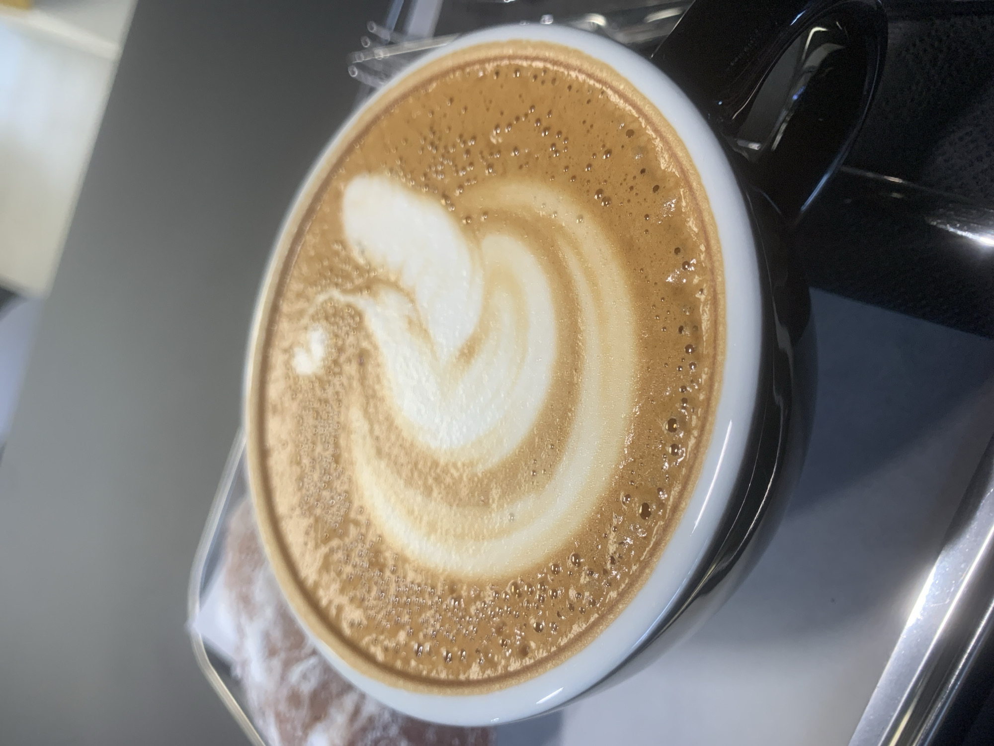 The image shows a close-up of a cappuccino in a black mug. The coffee has latte art, with a creamy white swirl in the center surrounded by a tan-colored foam. The mug is sitting on a metal surface, possibly a coffee machine or serving tray. The environment is well-lit. There is no discernible information to determine whether the image was taken in the city of Izhevsk, Russia.