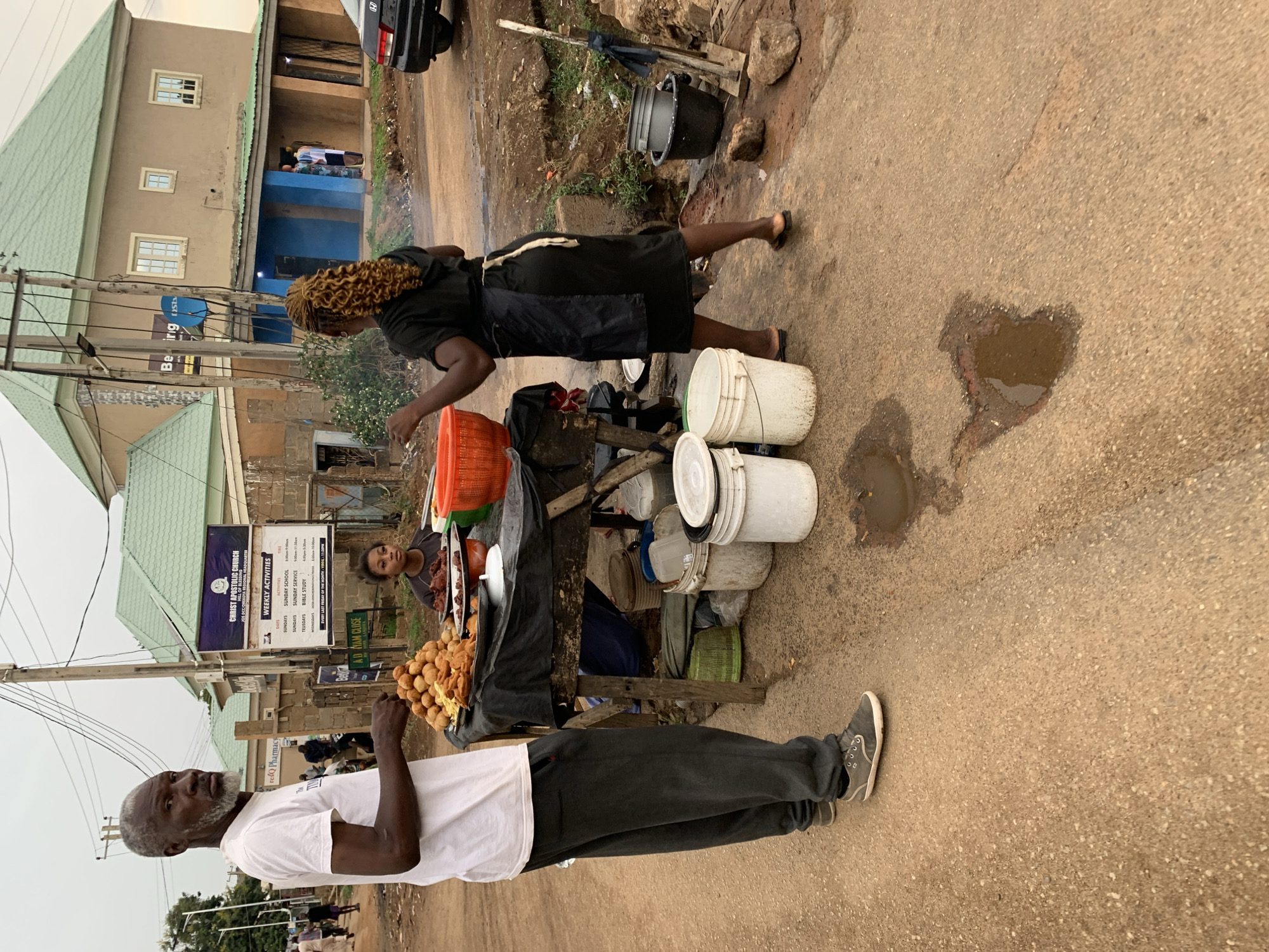 The image shows a street scene in New Stadium, Nigeria, featuring three people and a food stall. In the foreground, a man with a grey beard, wearing a white t-shirt and dark trousers, stands facing the camera. Beside him is a food stall laden with round, golden-brown snacks. A woman dressed in black is positioned behind the stall, appearing to be preparing or handling food items within an orange container. A young person can be seen behind the stall, partially obscured.
The background includes buildings, one with a green roof and another with a sign for Christ Apostolic Church and ResQ Pharmacy. There is also a black bucket on the ground and several white buckets on the stall. The road has puddles.