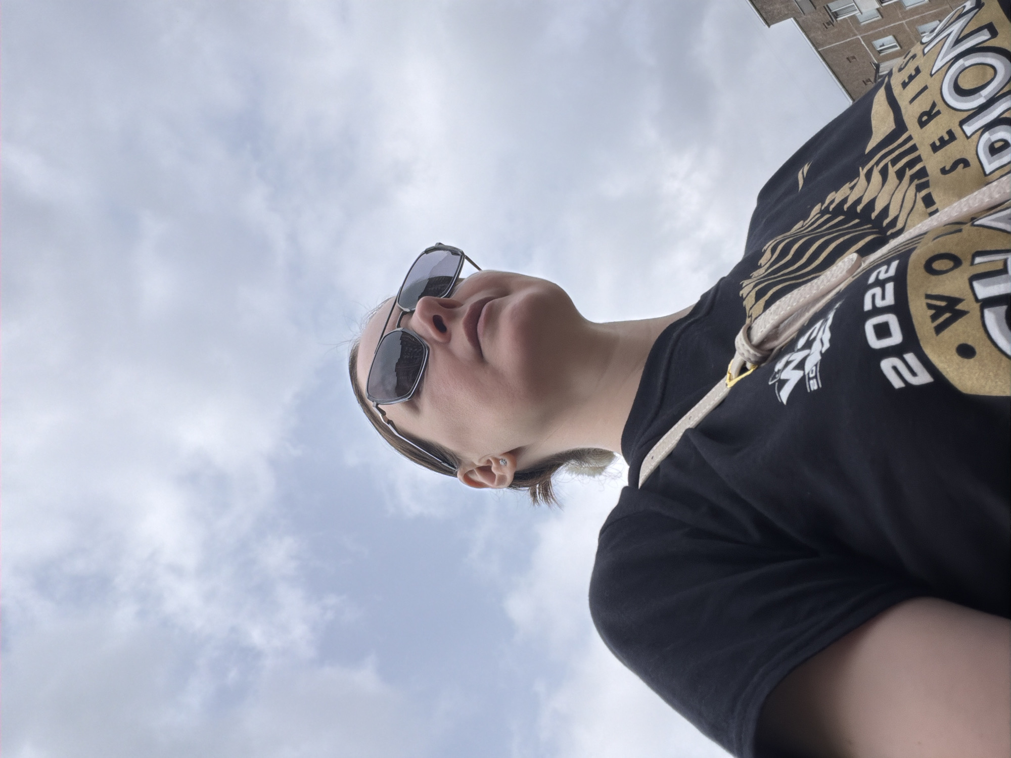 A low-angle photograph captures a person wearing sunglasses, a black t-shirt, and a bag with a rope-style strap. The t-shirt is emblazoned with "WORLD SERIES CHAMPIONS 2022" in gold and white lettering. The person looks up toward the sky, which is filled with light, wispy clouds. A building is partially visible in the top-right corner. The location is Unknown, Unknown.