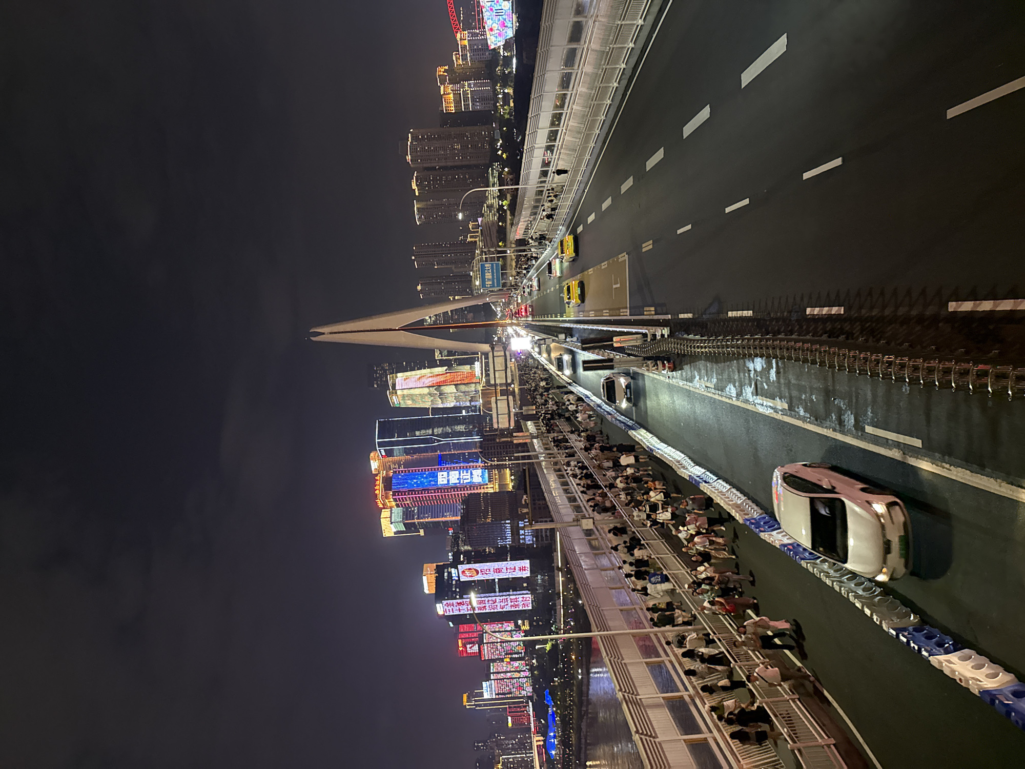 This is a night-time, eye-level, high-angle shot of a bustling bridge, likely the Hongyadong Bridge in Yuzhong District, Chongqing, China, given the iconic architecture and vibrant city lights. A wide bridge spans across a body of water, with a prominent, modern suspension tower visible on the left. The bridge has separate lanes for vehicles and pedestrians.
On the left side of the bridge, a pedestrian walkway is densely populated with people walking in both directions. They are dressed in casual attire, and the atmosphere appears lively. A white car is parked or moving slowly on the far left lane of the road, beside the pedestrian area. The roadway itself features multiple lanes of traffic. Several cars, including yellow taxis, are in motion on the right side of the bridge.
The background is dominated by a spectacular cityscape characterized by numerous tall buildings illuminated with colorful neon lights and digital advertisements. The sky is dark and cloudy, typical of a night scene. The bridge's structure, including railings and traffic barriers, is clearly visible, with wet patches on the road surface suggesting recent rain or the presence of water. The overall impression is one of a vibrant, urban night scene, with considerable human activity and a striking illuminated skyline.