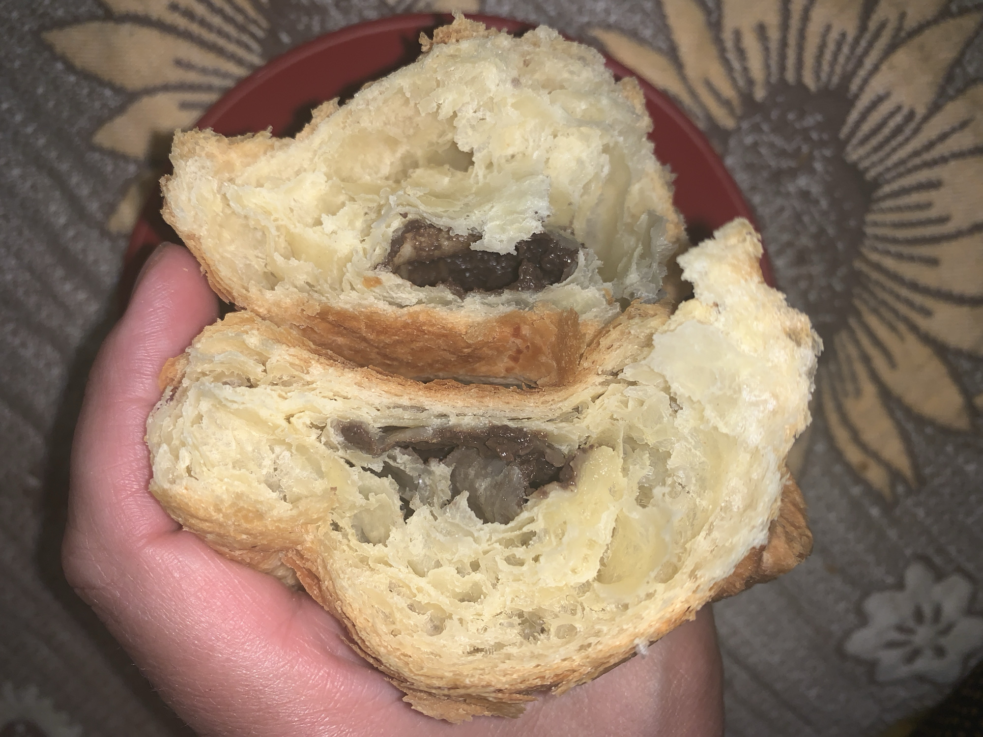 A close-up, top-down shot shows a hand holding a golden-brown, flaky pastry, which has been split in half to reveal its dark filling. The pastry exhibits distinct layers and air pockets typical of a croissant or similar viennoiserie. The interior filling is a rich, dark substance, possibly chocolate or a dark fruit paste, with a somewhat lumpy texture. The hand holding the pastry has visible skin texture and light hair on the fingers. In the background, parts of a plain red plate are visible, resting on a dark grey or brown patterned fabric. The fabric features lighter, stylized floral or sunburst patterns. The lighting highlights the texture of the pastry. No text is visible in the image.