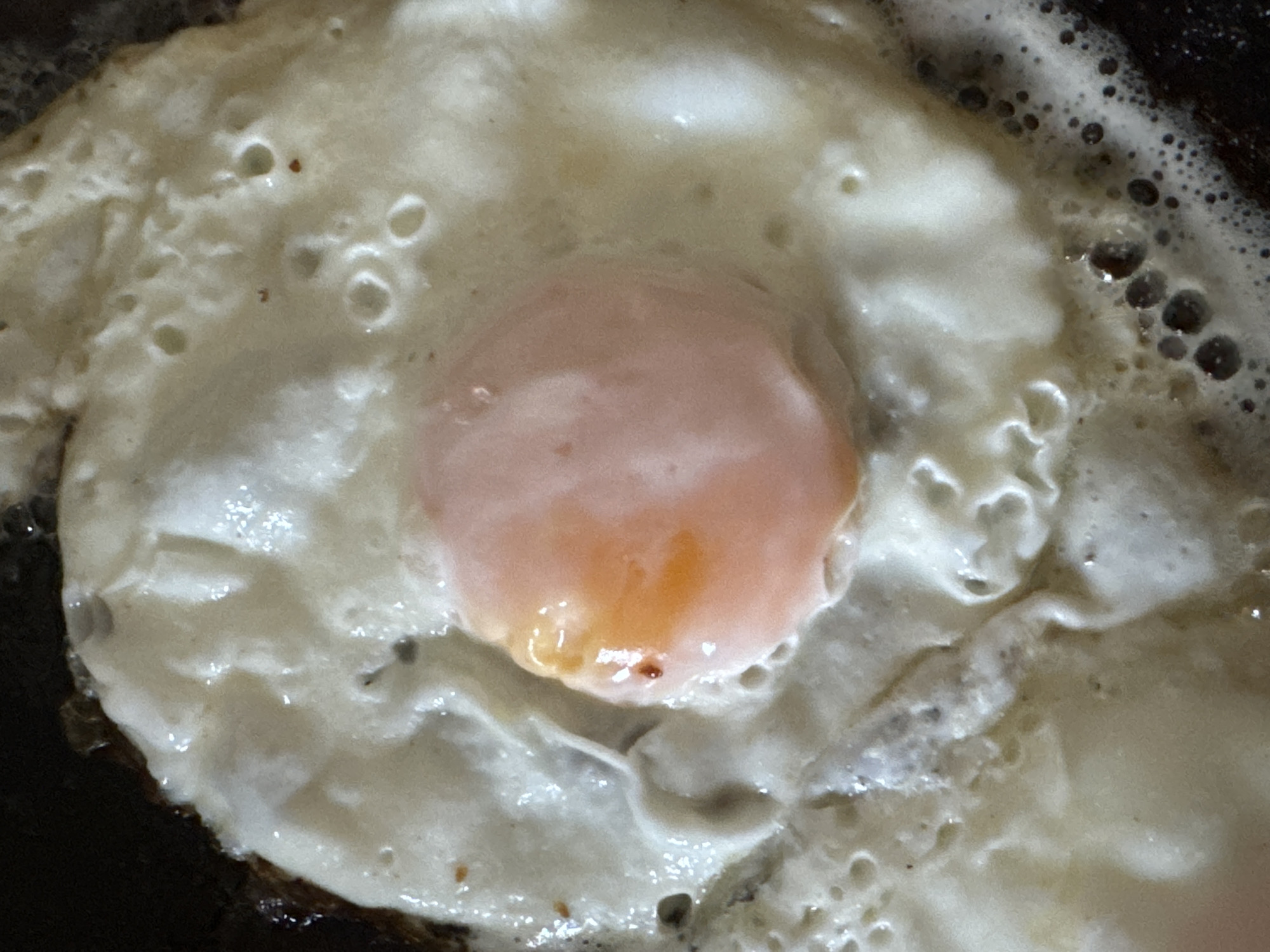 A close-up shows a fried egg with a runny yolk in a pan. The egg white is cooked and bubbly, while the yolk is pinkish and orange. Bubbles are visible around the edge of the egg, suggesting it is still cooking. The pan appears to be dark colored. The image does not contain enough information to determine a specific location in Chaikovsky, Russia.