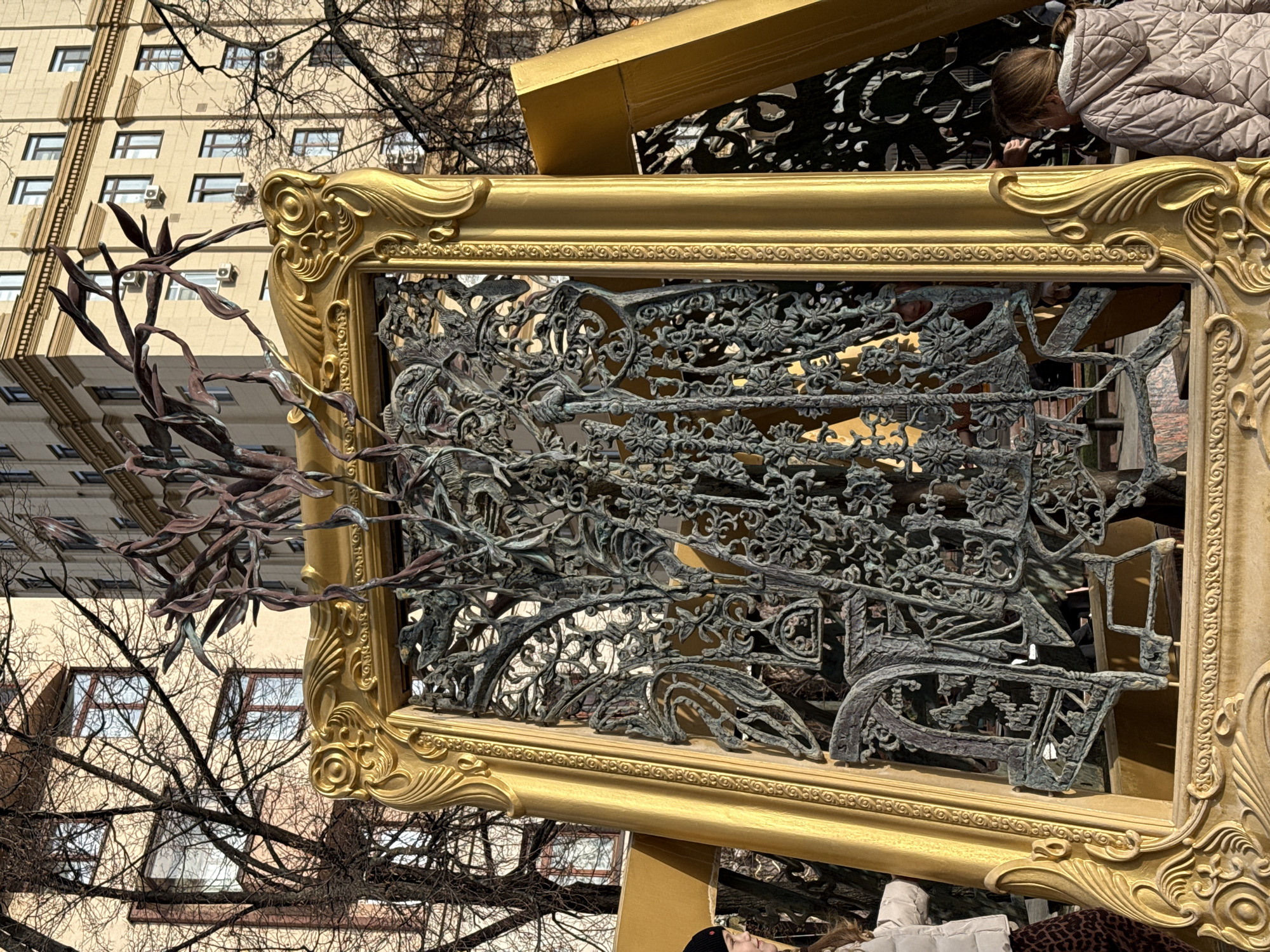 An outdoor, sunlit scene in the Patriarch's Ponds area of Moscow, Russia, features a prominent, large, gold-colored art installation. The central element is an ornate, rectangular frame, elaborately carved with classical motifs, standing upright and tilted slightly backward. Encased within this frame is an intricate, dark-patinated metal sculpture composed of dense, stylized organic forms resembling intertwining foliage, branches, and possibly obscured figures. Additional dark metal, leaf-like elements extend upwards from the top edge of the frame. To the right, a portion of a second, similar golden frame structure is visible, also tilted.
Two individuals are partially visible at the edges of the frame: on the left, a person wearing a black beanie and a light-colored jacket, looking towards the sculpture; on the right, a young girl with light brown hair tied back, dressed in a light grey quilted jacket, observing the installation from behind.
The background consists of bare deciduous trees with intricate branch networks and multi-story residential buildings. These buildings feature light beige facades and numerous rectangular windows. The scene is illuminated by bright natural sunlight under a clear blue sky, casting distinct shadows.