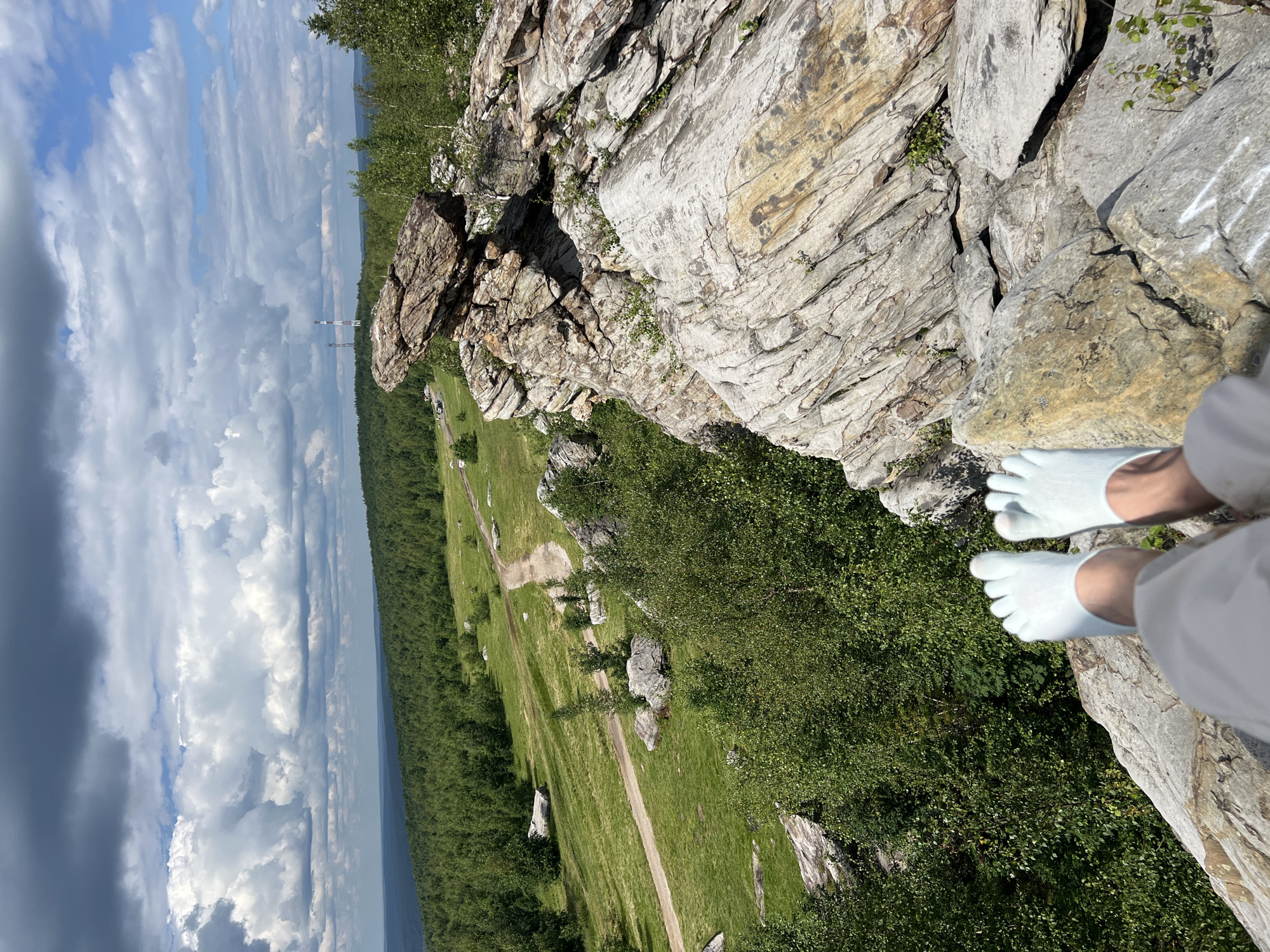 The image shows a person's feet, wearing light blue five-toe socks, dangling over the edge of a rocky outcrop. The person is seated on large, weathered rocks with a vast, green, forested landscape stretching out in the background. A dirt path winds through the grassy terrain. In the distance, two transmission towers are visible against a cloudy sky. This vantage point is from a rocky formation known as the Kliuchevskaya rock formation, located in the city of Gubakha, Russia. The weather appears to be partly cloudy, with a mix of sun and dark clouds. The foreground is dominated by the textured grey and brown rocks, with some sparse vegetation growing between them. The overall scene conveys a sense of elevation and natural beauty.