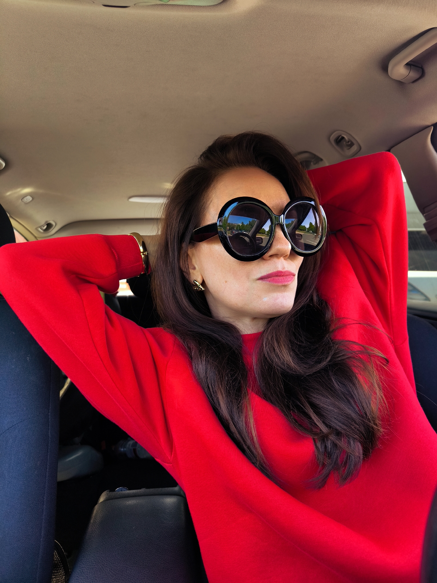 A woman with long brown hair is sitting inside a vehicle wearing large, round sunglasses and a red sweater. She has her hands behind her head. The reflection in her sunglasses shows trees, other vehicles, and buildings. Given the reflection in the sunglasses and the interior of the vehicle, the location is likely somewhere in the city of Riga, Latvia, based on general knowledge, but no specific landmark is evident to confirm a precise location. The car's interior is visible, including the dashboard, seats, and ceiling.