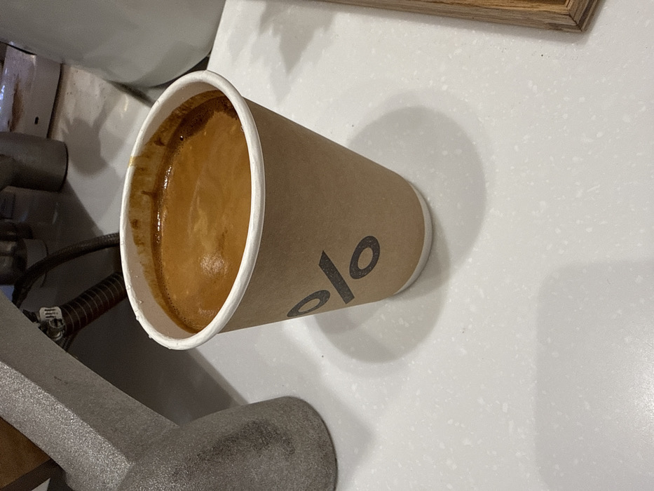 A disposable kraft paper coffee cup, marked with a prominent black percentage symbol (%) logo, rests on a speckled light gray countertop. The cup is filled to the brim with a dark brown liquid, topped by a thick, uniform layer of reddish-brown crema. To the upper left, a section of a textured, metallic gray object, consistent with a professional coffee machine component, is visible, featuring a dark hose or cable. A lighter, blurry surface occupies the upper right background. The scene depicts a prepared coffee beverage within a % Arabica coffee shop, located in Dubai, United Arab Emirates.