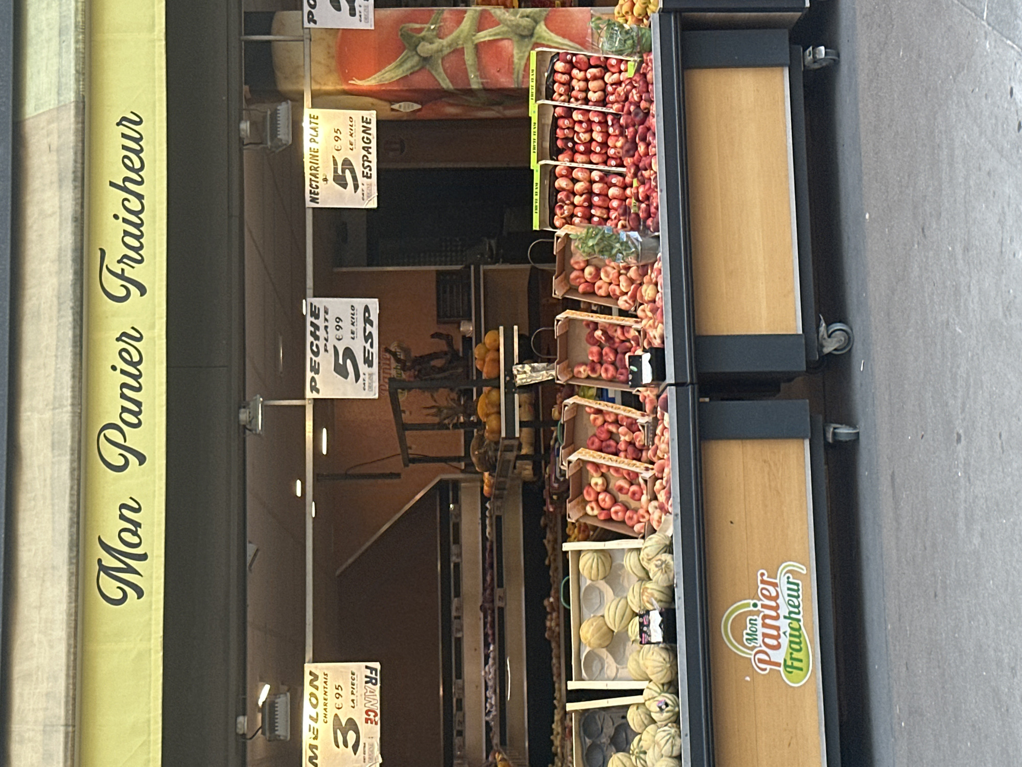 A frontal view captures a fruit stand in Paris, France, characterized by its yellow awning displaying "Mon Panier Fraîcheur" in cursive font. The stand is stocked with a vibrant assortment of fresh produce. Several price tags are visible, indicating items like "MELON CHARENTAIS" priced at €3.95 per piece, "PECHE PLATE" at €5.99 per kilo, and "NECTARINE PLATE" also at €5.95 per kilo, sourced from Spain. The produce is displayed in wooden crates and bins, featuring various types of melons, peaches, and nectarines. The background shows the interior of the shop with more fruits and vegetables arranged on shelves, along with lighting fixtures. The overall scene depicts a bustling market environment.