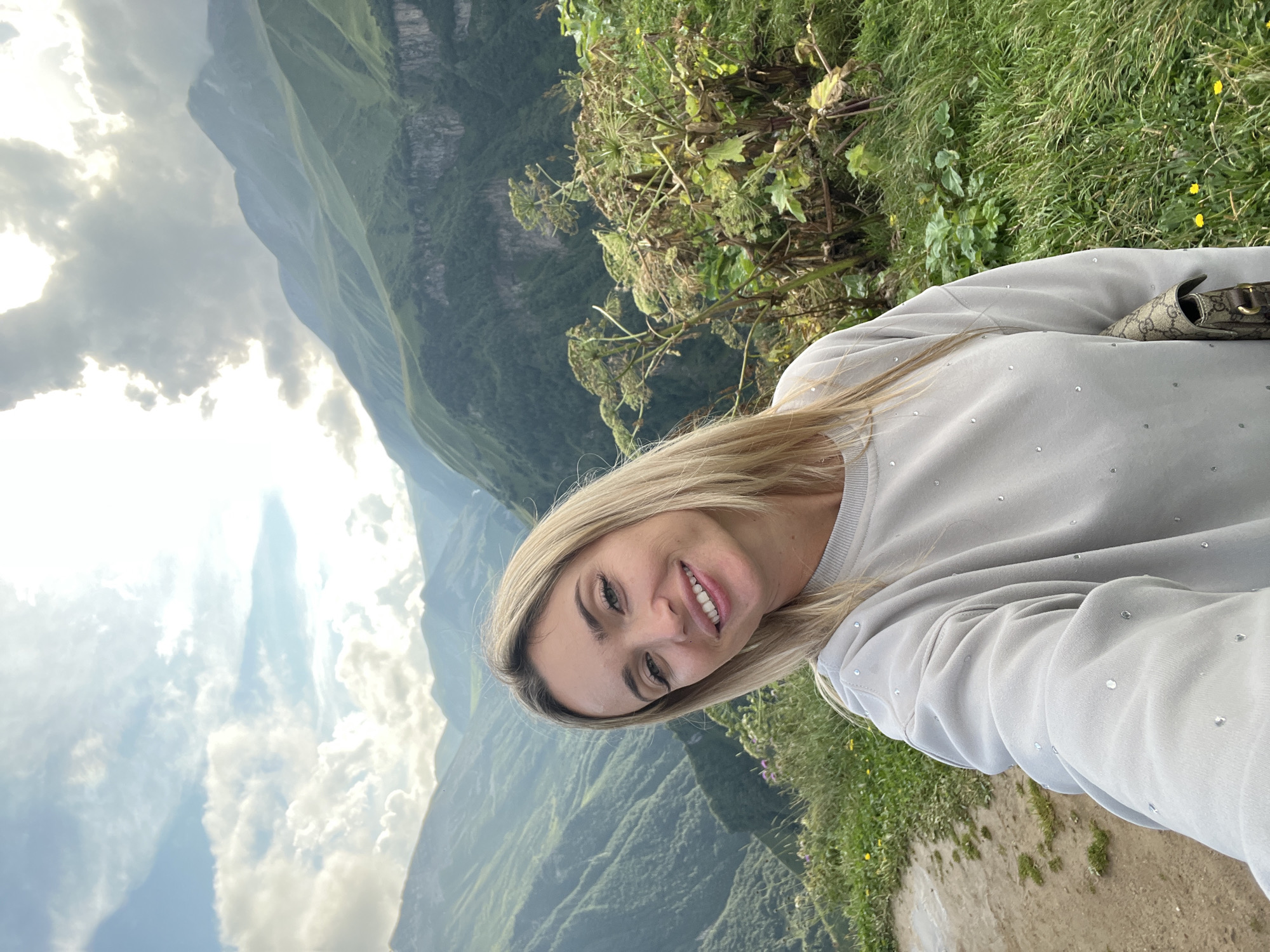 A blonde woman with fair skin and green eyes is taking a selfie on a mountaintop in Gudauri, Georgia. She is smiling and looking at the camera. The woman is wearing a light grey sweatshirt adorned with small, sparkling studs. She has a designer handbag slung over her shoulder. Behind her, a vast, rugged mountain landscape stretches out, characterized by steep, green slopes and dramatic rock formations. The sky above is a mix of bright sunlight breaking through clouds, casting a soft light on the scene. The foreground features lush green grass and some tall, leafy plants. The overall impression is one of a breathtaking natural vista combined with a personal moment of enjoyment.