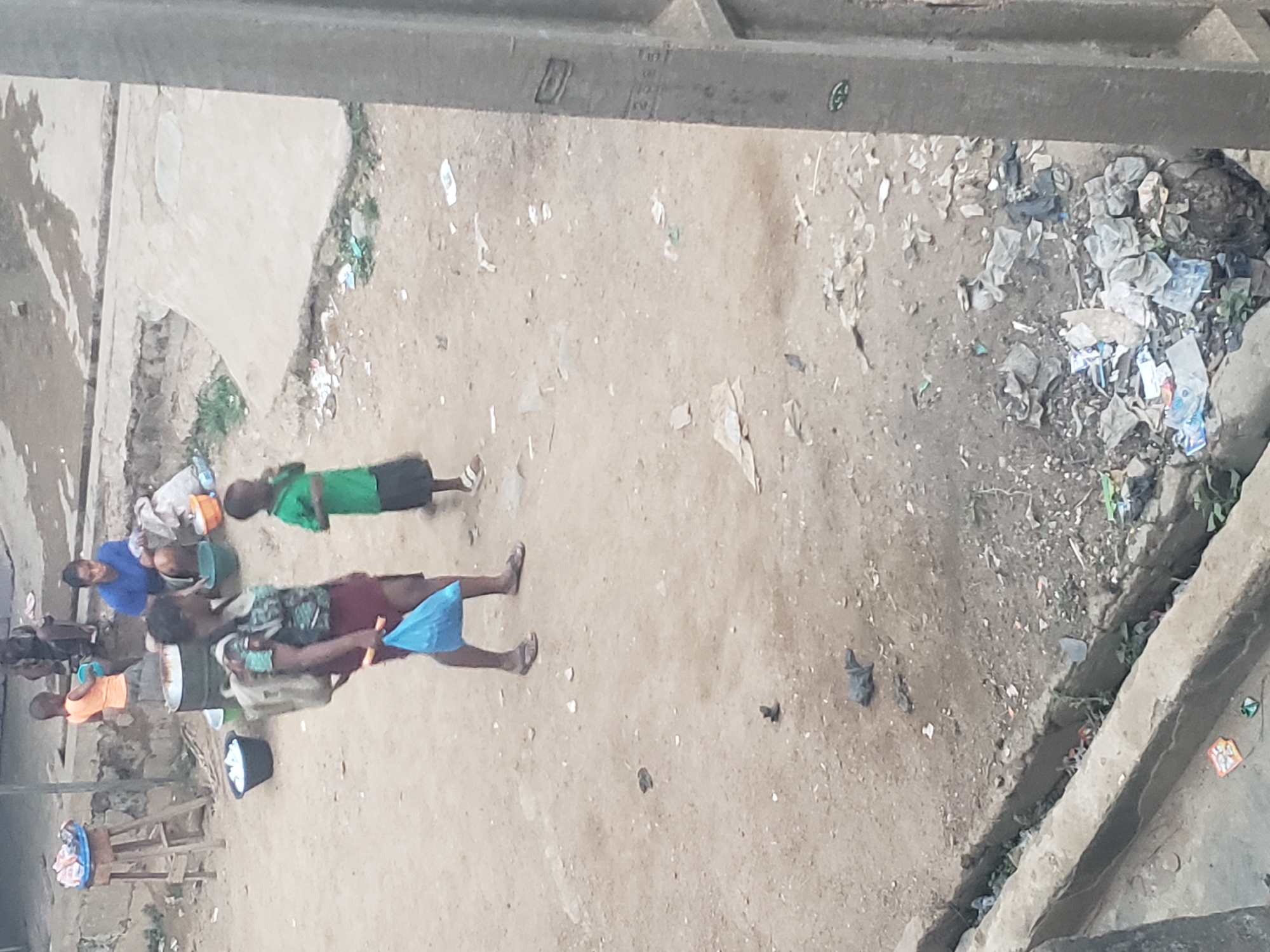 The image depicts an outdoor scene in Alalubosa, Nigeria. Several people are present, including children and adults. Some individuals are lying on the ground, while others are seated around what appears to be a cooking area with pots and containers. The surrounding environment is characterized by dirt and debris. There are some concrete structures visible in the background and along the edges of the scene. The location seems to be an open, unpaved area, potentially near a residential zone.
