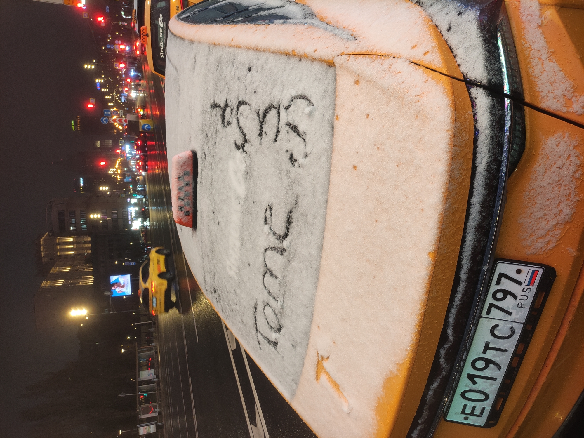 The image shows the back of a yellow taxi in Moscow, Russia at night. The taxi has a layer of snow on it, and the word "Susano" is written in the snow on the trunk. The license plate number is E019TC797, and the "RUS" indicator is visible on the plate. In the background, city lights are visible, along with other vehicles. The road appears wet from snow melting. The photograph is taken from a close, slightly elevated angle.