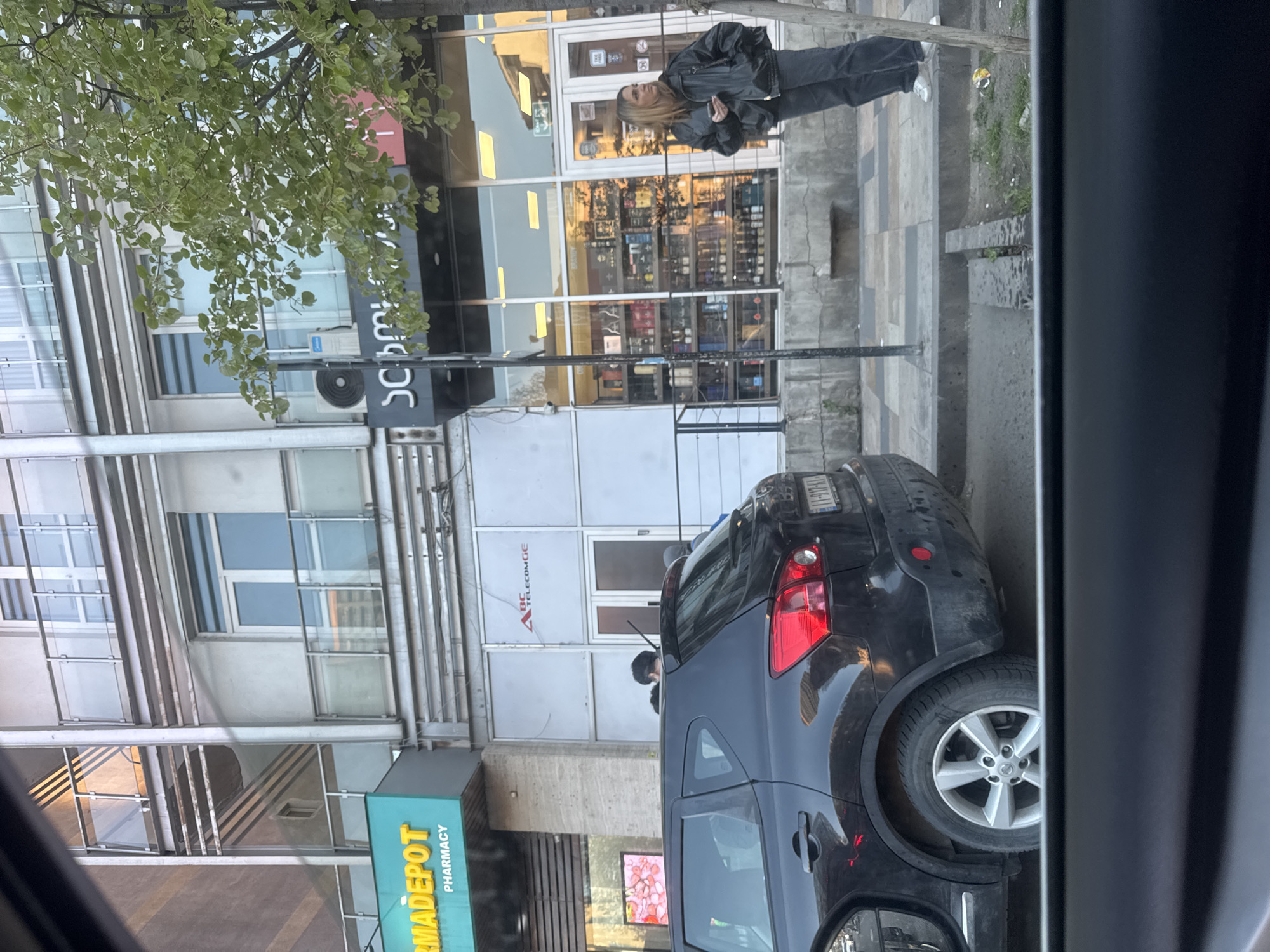 The image captures a rotated street view in Tbilisi, Georgia, seemingly from inside a parked vehicle during daytime, possibly under an overcast sky.
In the foreground, a black SUV is parked on the street, partially obscured by the frame and side mirror of the viewing vehicle. The SUV's rear wheel is prominent, and its Georgian license plate is partially visible as "I-077-AT". A person with dark hair is seen bending over near the rear of the SUV.
On the opposite side of the street, several commercial buildings line the pavement. A large green tree with light green leaves hangs over the scene, partially obscuring the buildings.
A woman with long blonde hair, wearing a dark jacket and trousers, stands on the paved sidewalk, looking towards the left. She appears to be holding a small dark object. Behind her is a storefront with large glass windows reflecting various items inside, suggesting a retail business. Above this store, a dark sign with partially visible white text in Georgian script, including "SCH," is present.
Further down the street, another storefront displays a light blue-green sign that reads "PHARMADEPOT PHARMACY". Additionally, a sign for "BC TELECOM GE" with a red triangular logo can be seen on another building. The pavement consists of grey concrete slabs, with some visible dirt and sparse vegetation. The scene depicts a typical urban environment.