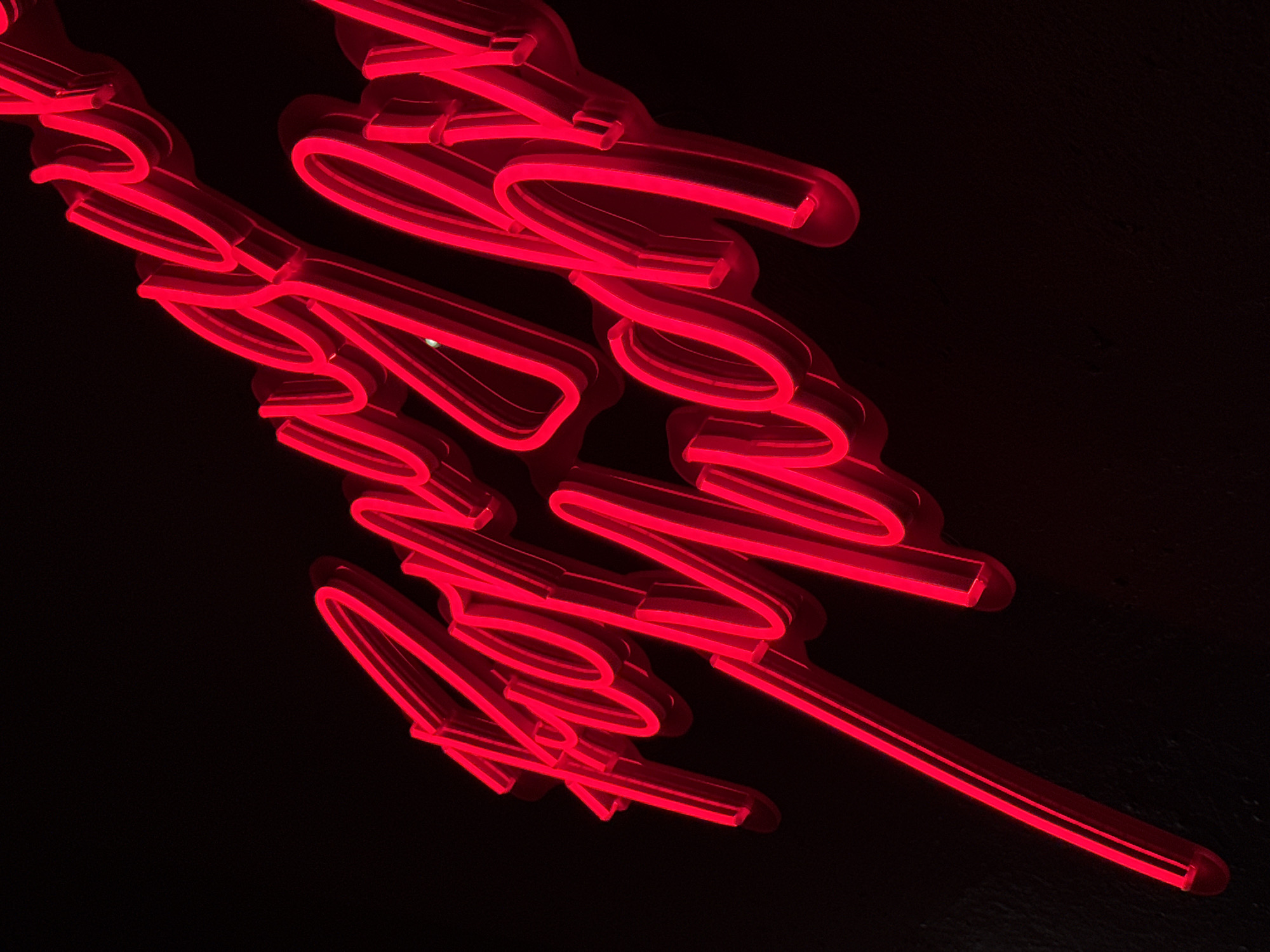 This media file features red neon lights against a black background. The neon lights form cursive lettering, creating a dramatic and somewhat abstract visual. The lighting is intense, with the red glow illuminating the shapes of the letters and casting subtle shadows. The overall impression is one of modern artistry or a stylish interior design element. The composition is dynamic, with the neon script flowing diagonally across the frame. There are no people, animals, or clear environmental cues to suggest a specific time of day, weather, or location beyond what can be inferred from the artificial lighting. The focus is solely on the glowing red text, which appears to be a word or phrase rendered in a decorative font. The texture of the black background is visible, suggesting a matte surface that absorbs light. The image evokes a sense of excitement, nightlife, or perhaps a sophisticated brand identity, depending on the implied meaning of the text.