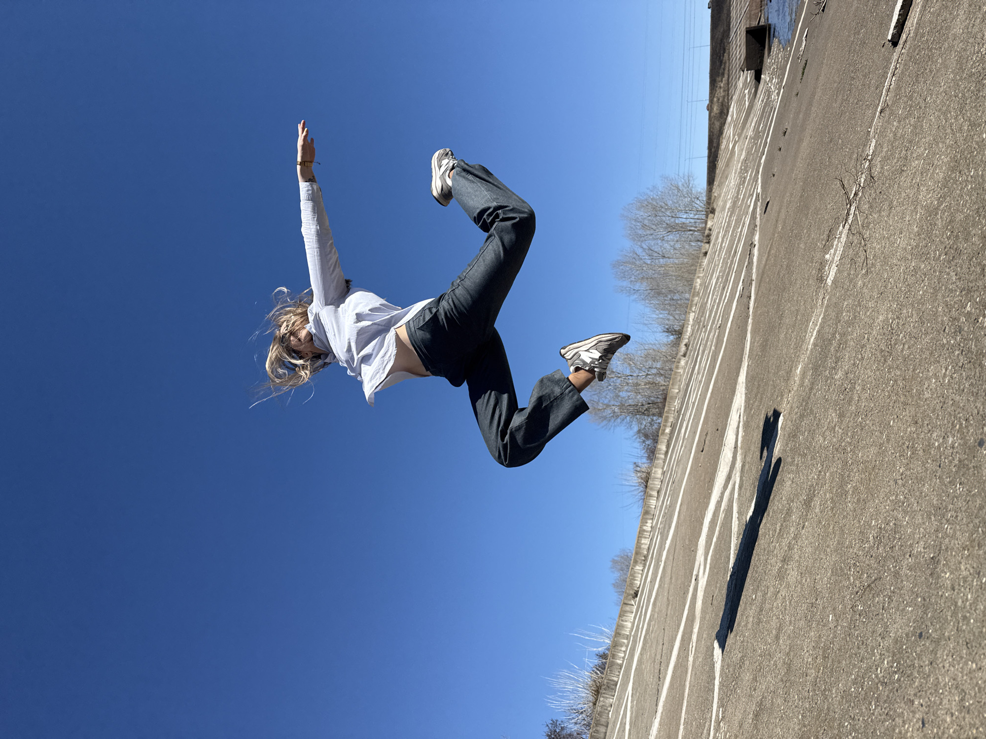 A young woman with blonde hair is captured mid-air, appearing to jump or leap with her arms and legs extended. She is wearing a light-colored button-down shirt, dark pants, and athletic shoes. The background consists of a clear blue sky above and a concrete structure below, which features white painted lines. The environment suggests an urban or industrial setting. The shadow of a person is cast on the concrete wall. The location is identified as Chaikovsky, Russia, based on available information.