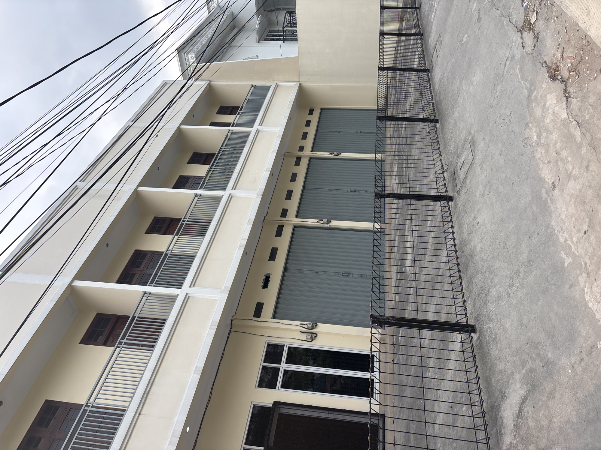 The image shows a two-story building with a beige facade and dark brown wooden windows. The upper floor has balconies with metal railings. The ground floor features several large gray roll-up doors, suggesting it is a commercial space or garage. A black metal fence runs across the front of the property, separating it from a concrete area that appears to be a sidewalk or driveway. Overhead, a complex network of black electrical wires crisscrosses the sky, obscuring the cloudy weather. The building is located in a dense urban environment, as indicated by the surrounding structures and infrastructure. Based on the architectural style and the presence of overhead electrical wires, this scene is consistent with many commercial areas in Bekasi, Indonesia. There are no people visible in the image.