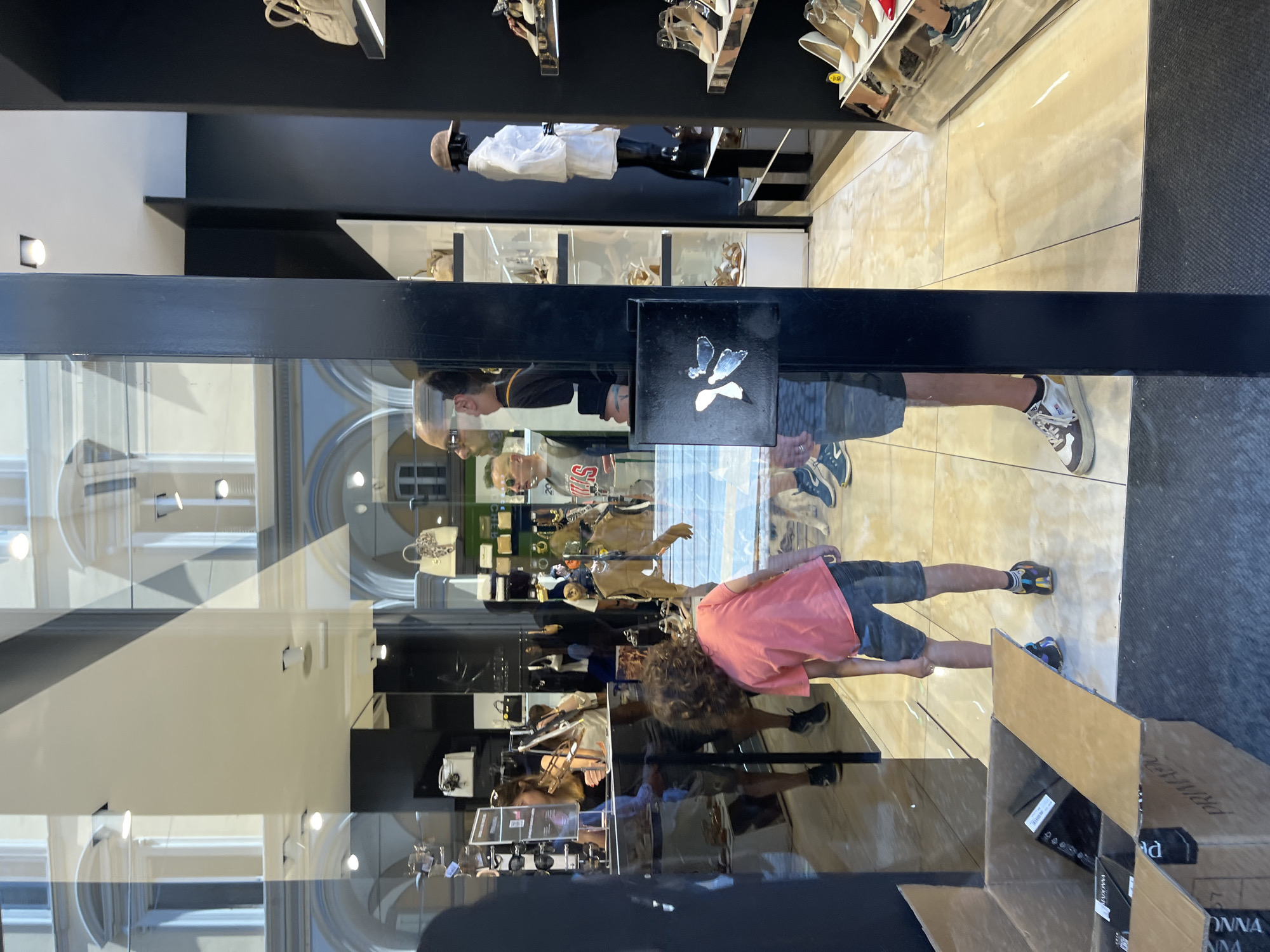 The image shows the storefront of a retail store in Bologna, Italy, reflected in a glass window. Inside, shelves display handbags and shoes. A mannequin stands near the top shelf. The reflections on the glass show two people, likely a child in a pink shirt and an adult, standing outside the store. Several cardboard boxes sit on the ground near the bottom right of the window. The reflection shows architectural details, including arched windows, of buildings across the street.