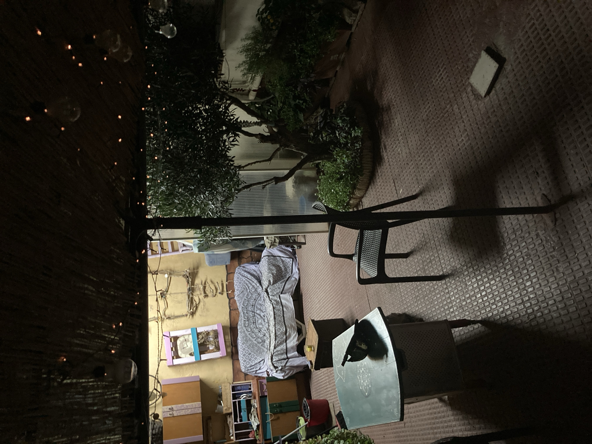 The image depicts a dimly lit outdoor patio area, likely in Sabadell, Spain, at night. A thatched roof covers part of the space, adorned with string lights and a few exposed bulbs. Beneath the roof, a couch draped with a patterned cover faces a wall with shelving and decorative items. To the left, a green metal table with a dark object on it, possibly a bag, stands in the foreground. A black plastic chair is positioned near the table. To the right, a small olive tree grows within a circular planter surrounded by lush greenery. In the background, more plants in planters are visible. The ground is covered in a red, textured material. A square, light-colored tile or object lies on the ground to the right of the frame.