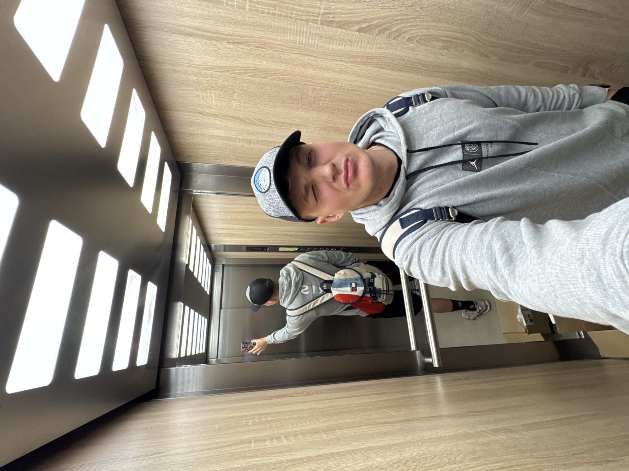 The image captures a young man taking a selfie inside a modern elevator, situated in Plovdiv, Bulgaria. He is looking directly at the camera with a playful wink or squint and a slight smirk. He wears a grey hooded sweatshirt featuring dark blue or black stripes on the shoulders, and logos for "PSG" (Paris Saint-Germain) and Jordan Brand are visible on the front. A grey baseball cap with a blue and white mountain-like emblem completes his casual attire.
The elevator's interior walls display a light wood grain pattern on the upper and lower sections, while the side panels are a sleek dark grey or silver. Bright, rectangular light fixtures are integrated into the left wall, and their reflections are visible on the opposite side. The floor also features a matching wood pattern.
A clear reflection of the man is visible in the shiny elevator wall directly in front of him. In the reflection, he is seen from behind, pressing an elevator button with his right hand. He is wearing a red, white, and blue sling bag or small backpack, with "TOMMY HILFIGER" prominently displayed on it. The scene is well-lit, conveying a modern and clean environment. The overall mood is casual and relaxed, indicative of an everyday moment.