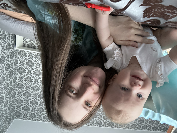 A woman with long brown hair and a baby are posing for a selfie. The baby is wearing a white ruffled onesie and has blue eyes. The woman has light eyes and is wearing a dark shirt. The background consists of patterned wallpaper and a patterned pillow. The woman is holding a red object above the baby.