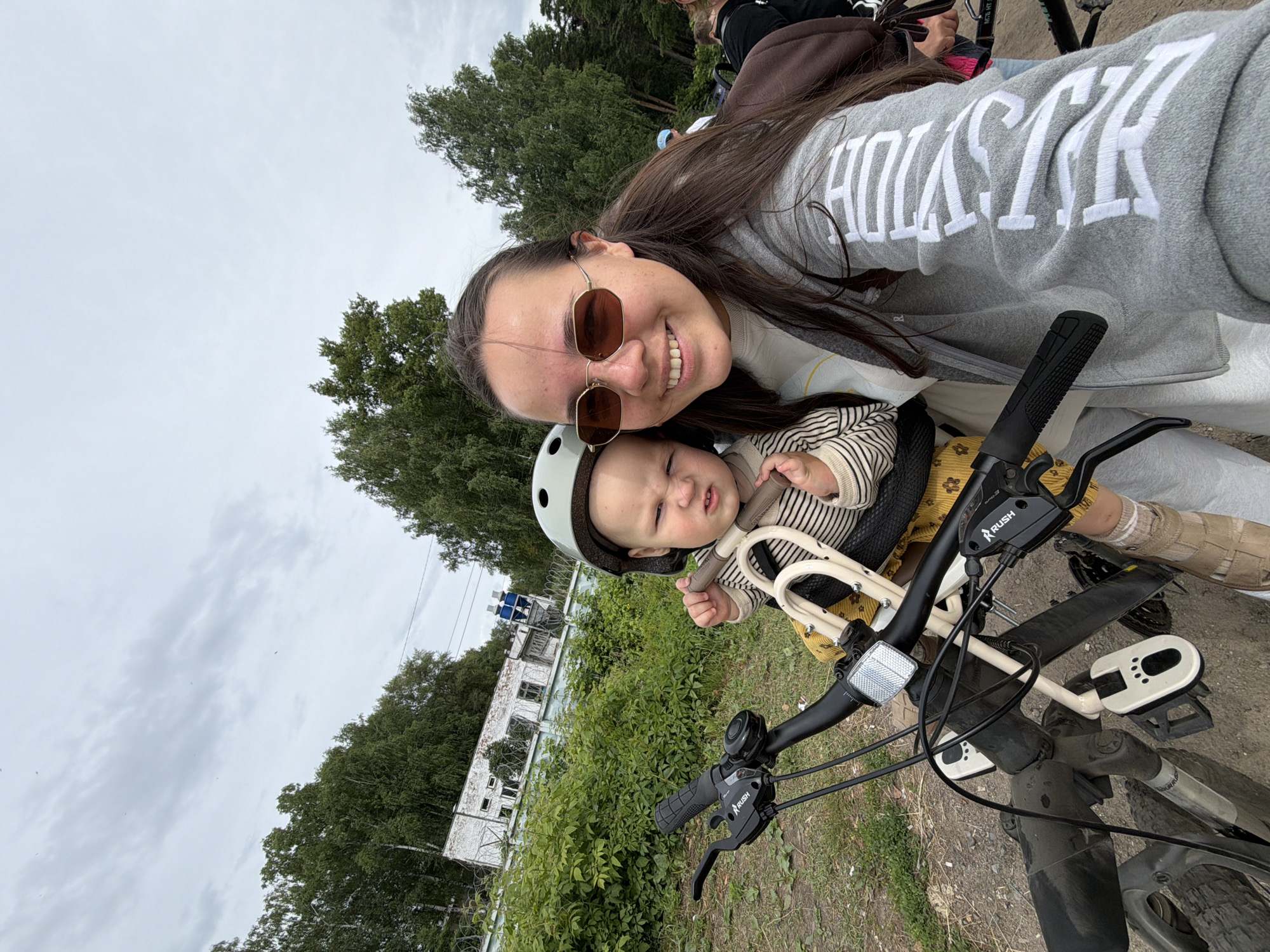 In Chaikovsky, Russia, a young woman with long dark hair and sunglasses is taking a selfie from a bicycle. She is smiling at the camera. A baby wearing a helmet is seated in a child seat on the front of the bicycle, facing forward and looking at the camera with a displeased expression. The bicycle is dark in color and appears to be in a dusty outdoor setting. In the background, there is a building with a fence and lush green trees, suggesting a semi-urban or park-like environment. The sky is overcast with some clouds.