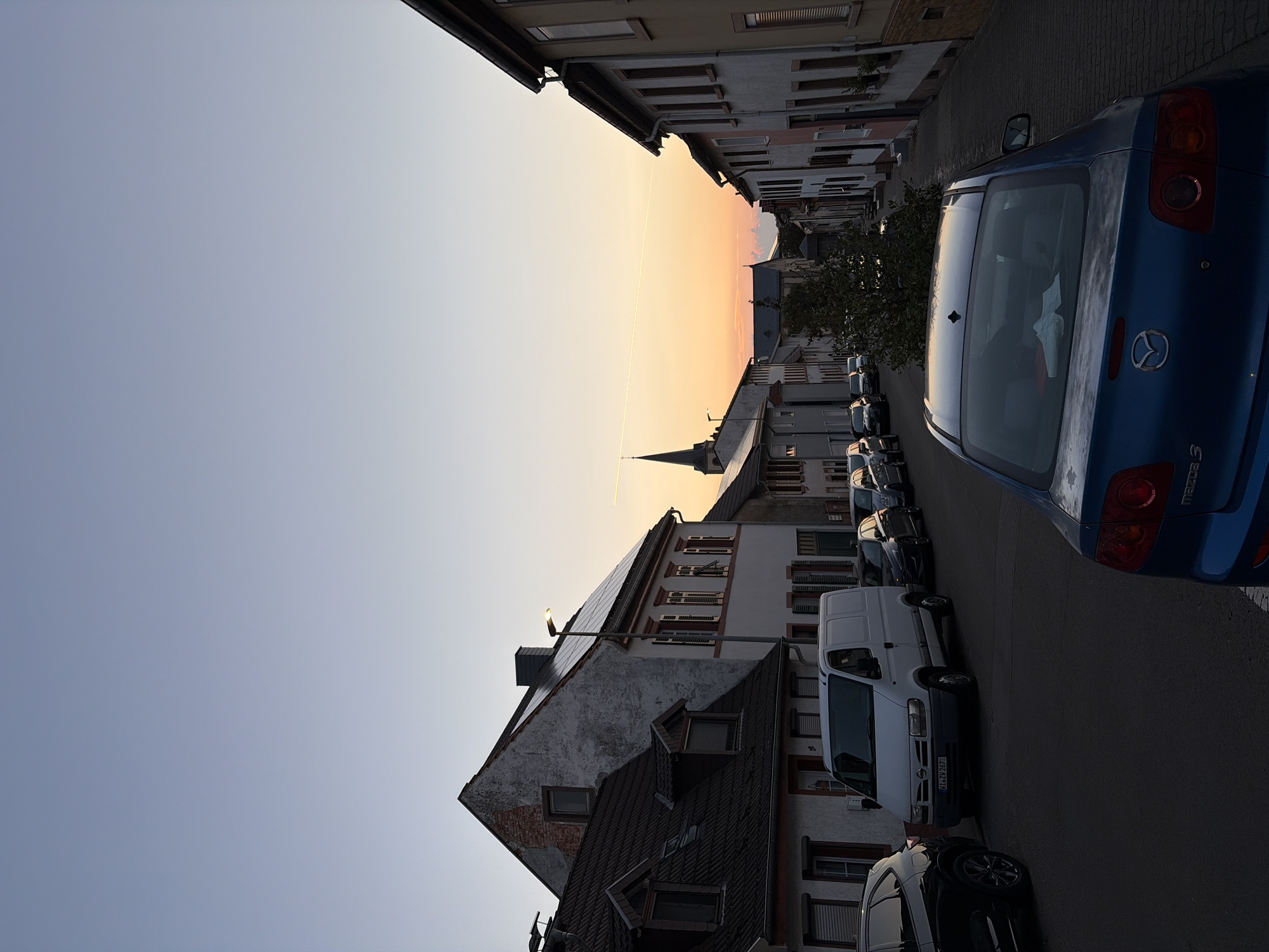 This is an eye-level, medium shot of a street scene in Kirchheimbolanden, Germany, during twilight. The sky is a gradient of pale blue to soft orange, with a faint contrail visible. The street is paved with cobblestones and lined with multi-story buildings featuring traditional German architecture with tiled roofs, shutters, and windows.
Several cars are parked along the street. In the foreground, the rear of a blue Mazda 3 is visible. To the left, a white Nissan van is parked, followed by several other cars. A black SUV is parked on the far left.
In the background, a church steeple with a cross is visible, indicating the presence of a church. A small, leafy tree is situated between buildings in the middle distance. The overall atmosphere is calm and serene, characteristic of a quiet evening in a European town. The image appears to have been taken from a slightly elevated position looking down the street.