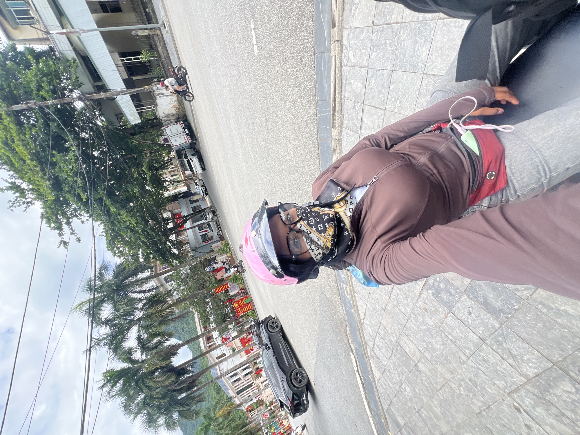 A person wearing a pink helmet, glasses, and a brown jacket is sitting on a scooter, facing the camera. They have a red fanny pack around their waist, with a phone and earbuds visible. The scooter is on a paved sidewalk next to a street. Across the street, there are buildings, palm trees, and a black SUV. In the background, there are more trees, buildings, and a cloudy sky. The image appears to have been taken in the city of Hà Giang 2 Ward, Vietnam.