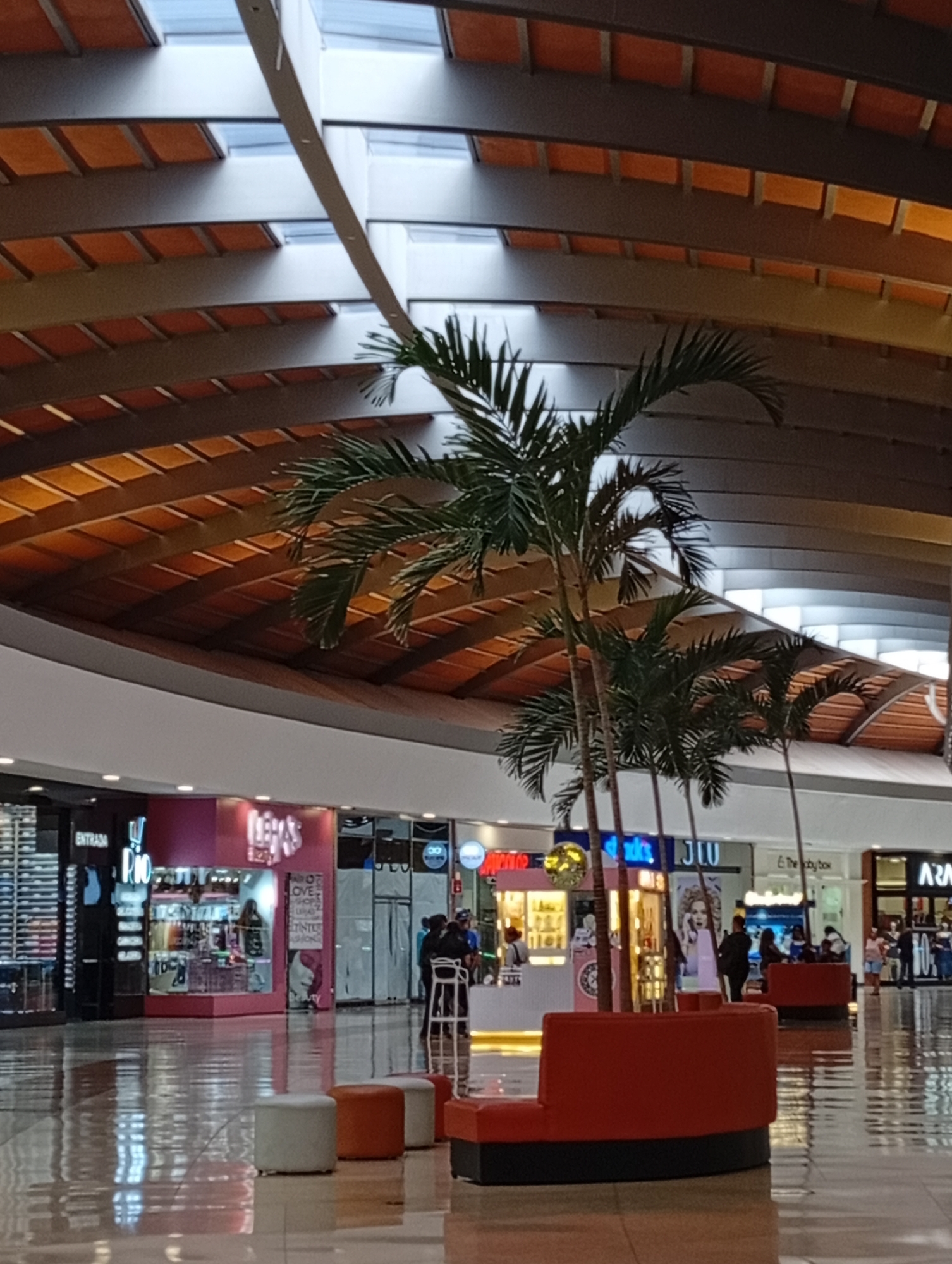 The image captures the brightly lit interior of a modern shopping mall. A high, arched ceiling, characterized by dark beams and light wooden paneling, features multiple skylights that allow natural light to filter in. Several tall, slender palm trees are arranged along the central walkway, serving as decorative elements and separating the main path from seating areas.
In the foreground, a seating arrangement consists of two curved red benches with black bases, alongside smaller cylindrical ottomans in white and orange. The highly polished floor reflects the overhead lighting and the storefronts, creating a glossy sheen.
Along the sides of the mall, various retail stores are visible with brightly lit displays and facades. Several people can be seen in the background, walking, standing, or browsing inside the stores. Their activities suggest a typical shopping environment. One person is seated on a stool near a counter.
Visible text on store facades includes "ENTRADA" (Entrance) and "RIO" on a black and pink storefront to the left. Another pink store displays "LEKAS" and lists items like "LOVE", "SHOP", "ZUMBA", and "TINTES" on its window. Further down, store names like "JIU", "E The Joy Box", and "ARA" are discernible.
The scene conveys a spacious, clean, and inviting commercial atmosphere, likely during the daytime due to the presence of natural light from the skylights, combined with indoor artificial lighting. The setting is consistent with a contemporary shopping center in Pampatar, Venezuela.
